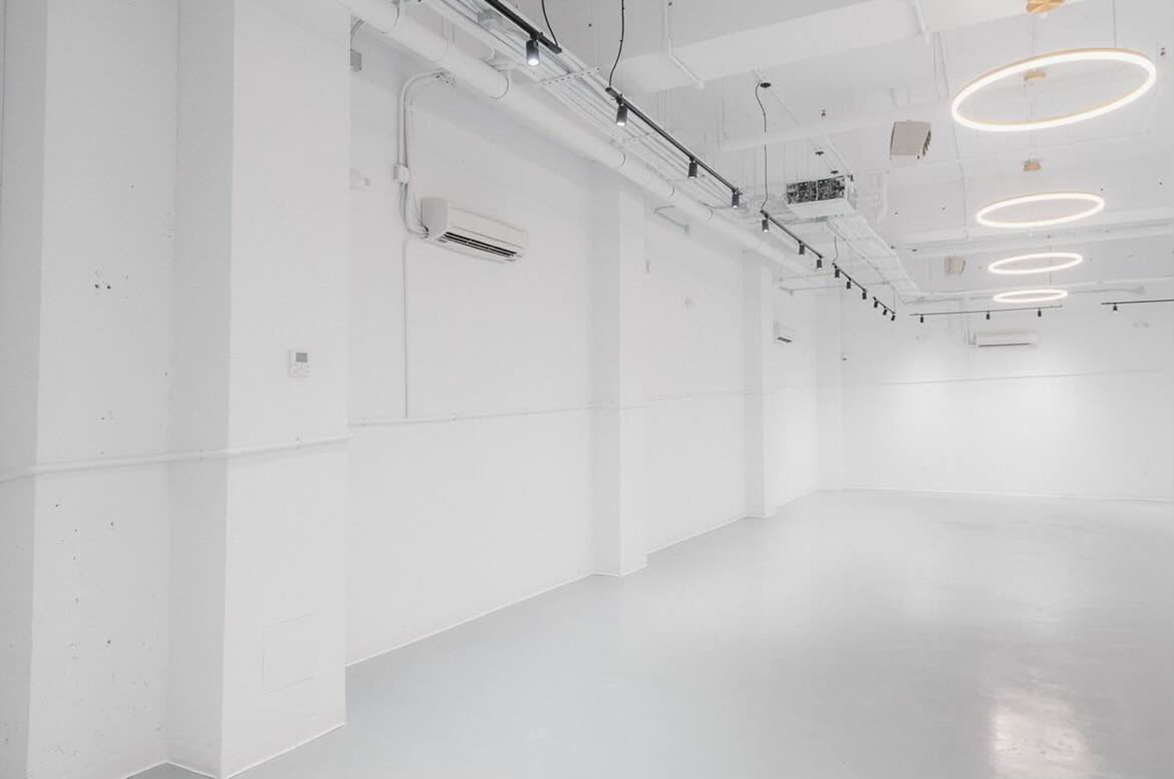 The image shows a spacious, minimally decorated room with white walls, a polished floor, and circular ceiling lights.