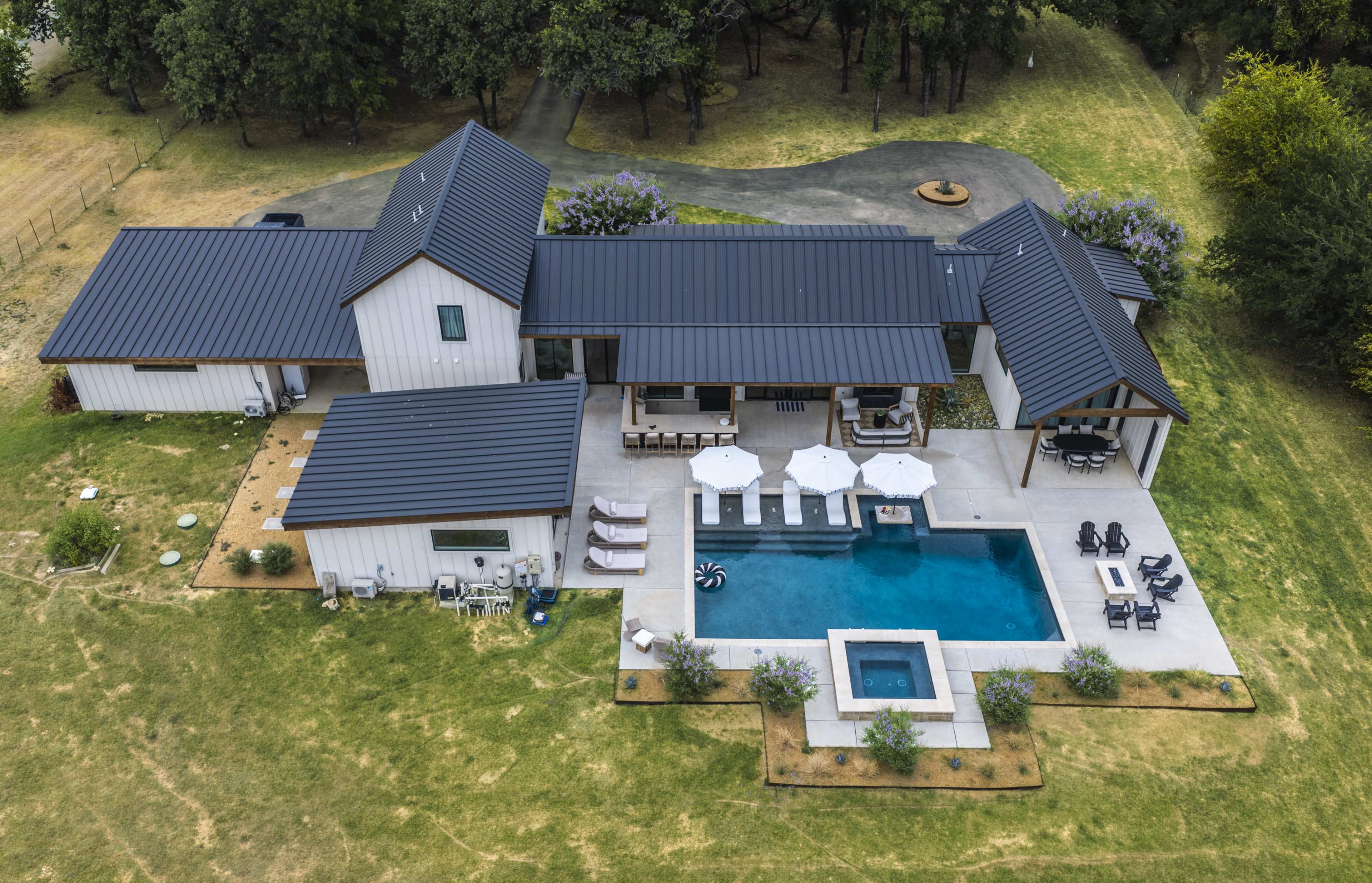 The image shows a modern single-story house with a dark metal roof, a swimming pool, and a patio area surrounded by landscaping.