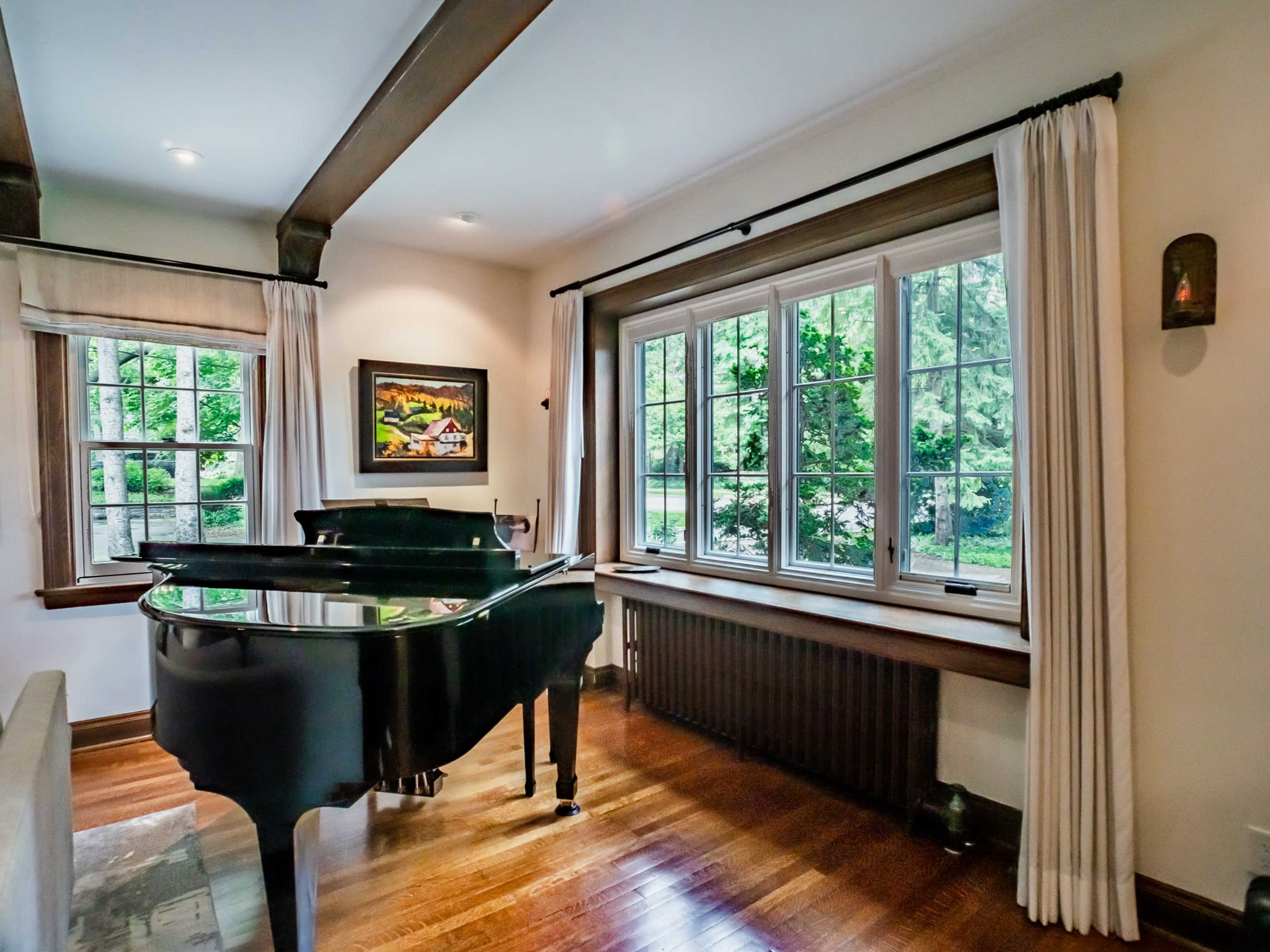 A black grand piano is positioned in a well-lit corner of a room with large windows and wooden trim, overlooking a green outdoor space.