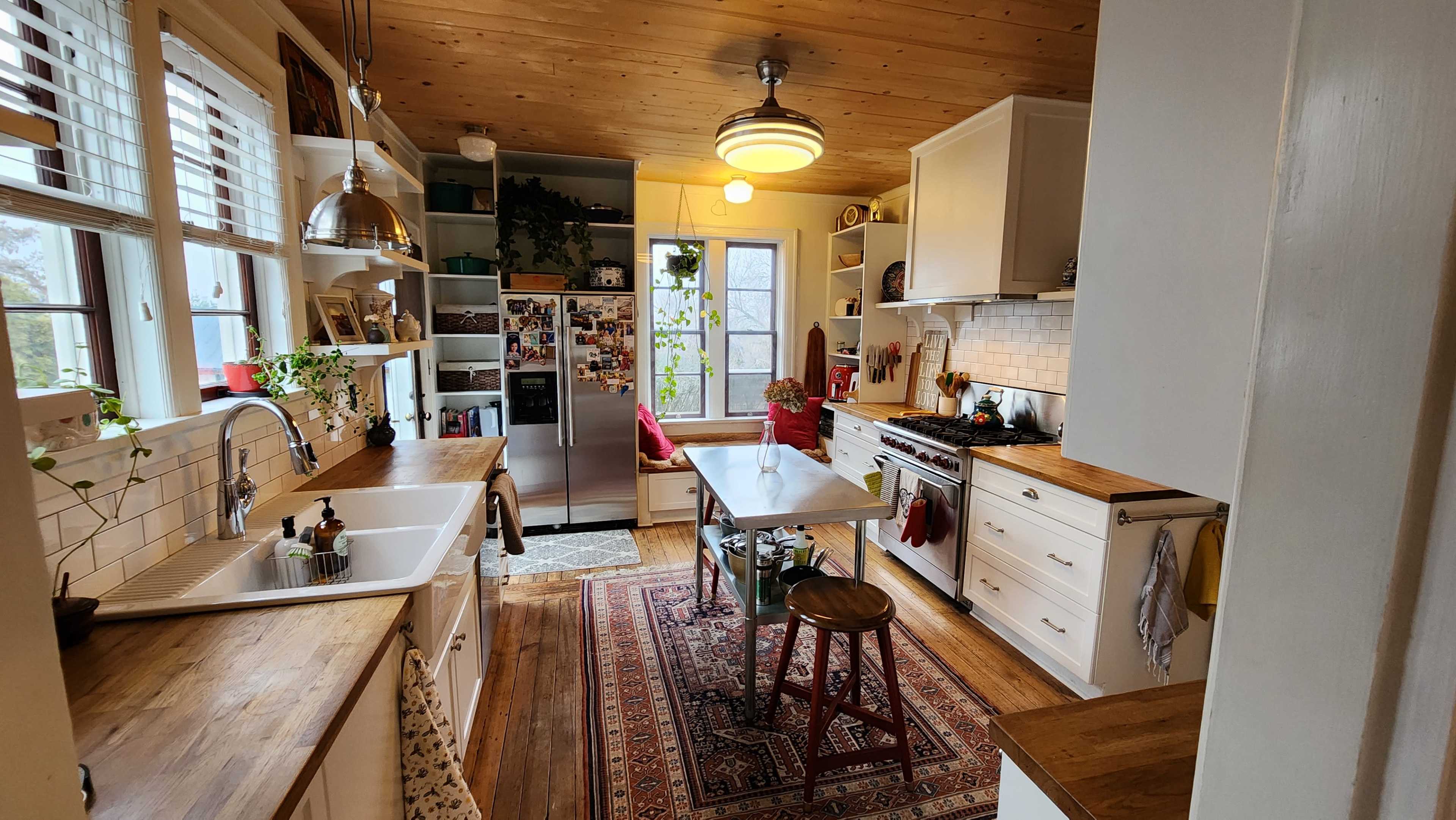 A bright kitchen features wooden cabinetry, a central table with a stool, and a mix of modern appliances and decorative plants.