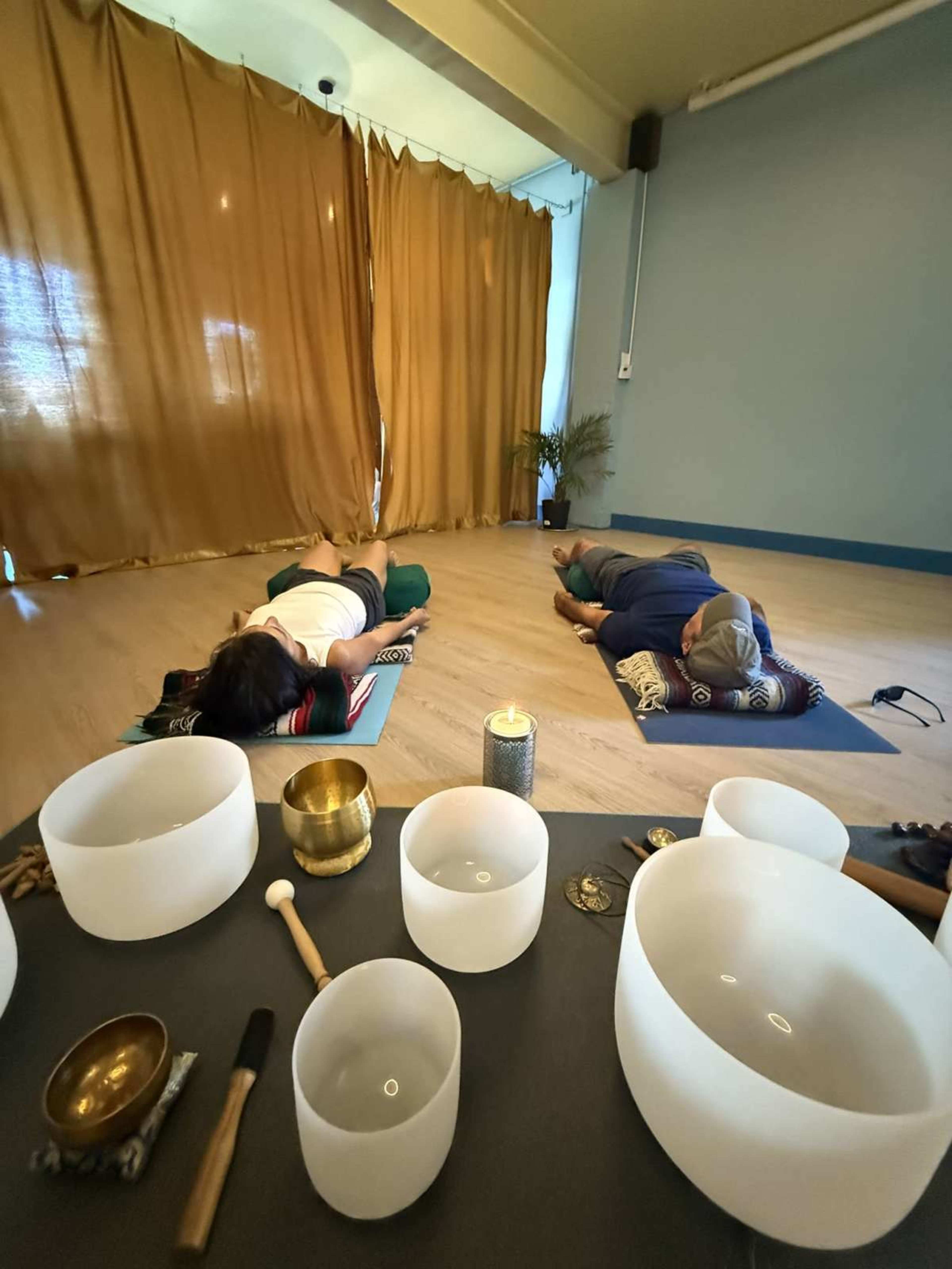 Two individuals lie on yoga mats in a serene space adorned with golden drapes and various meditation instruments in the foreground.