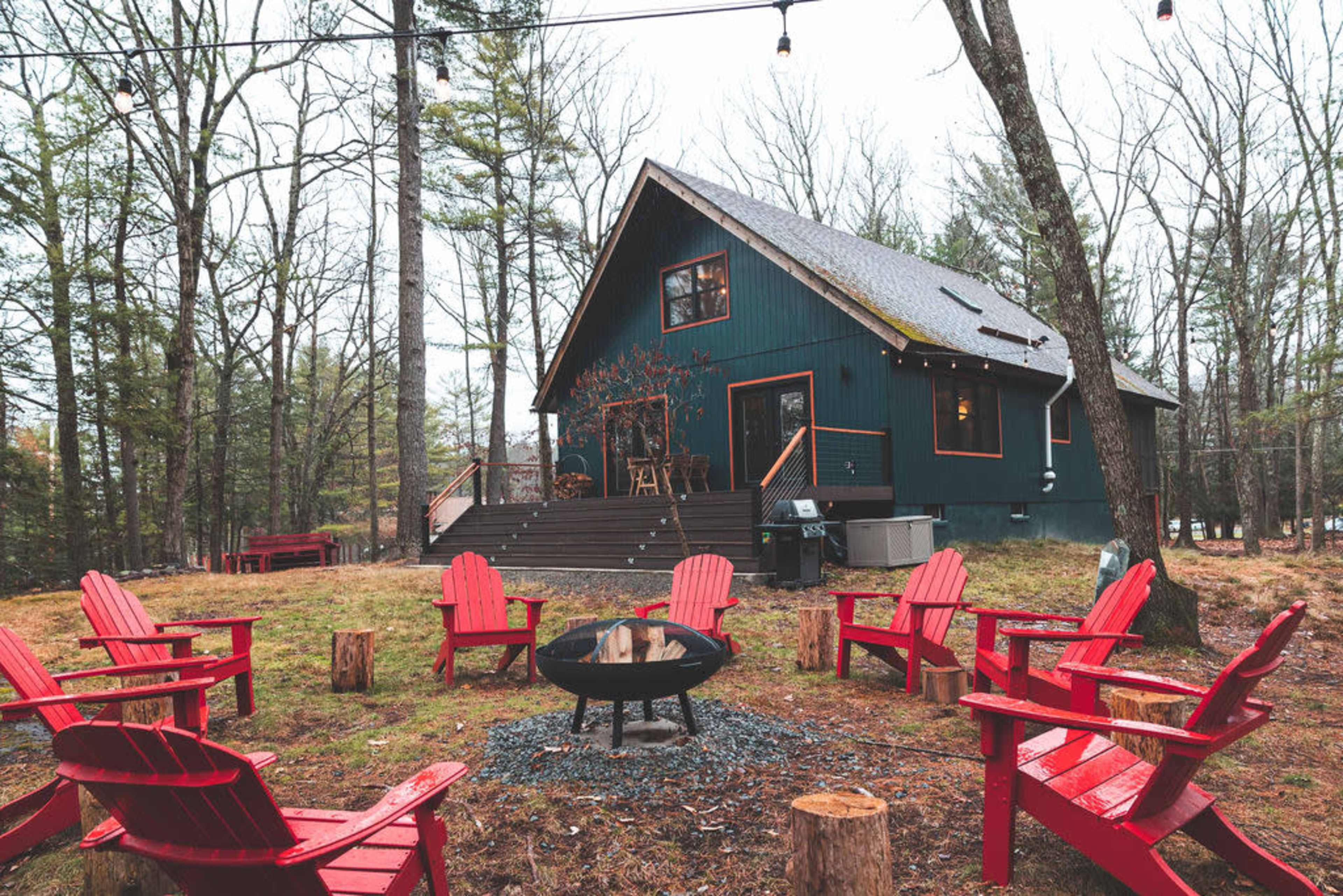 A green two-story cabin is surrounded by red Adirondack chairs and a fire pit in a wooded area.