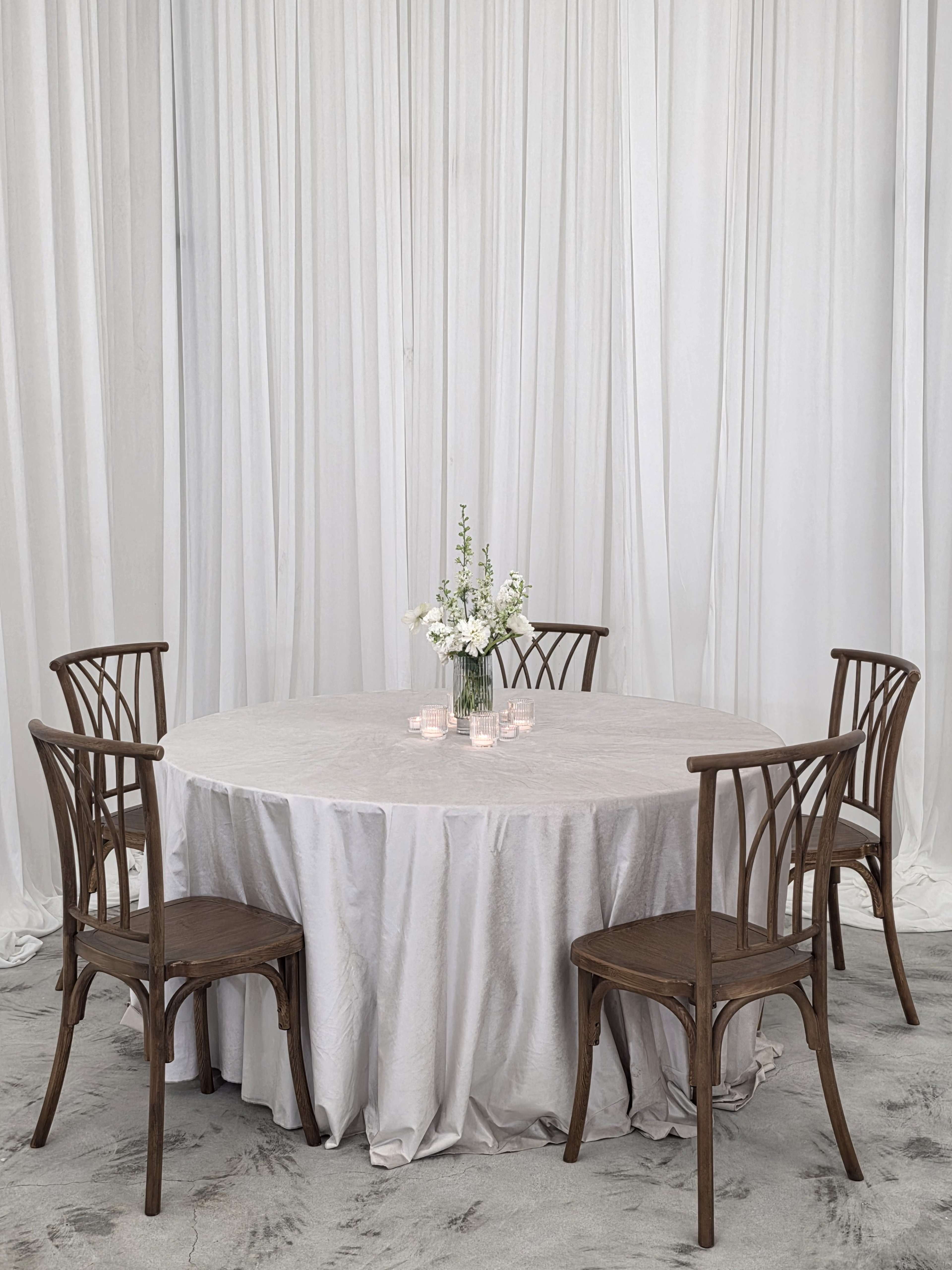 A round table covered with a light-colored tablecloth is set with a floral centerpiece and surrounded by four wooden chairs in a space with white drapery.