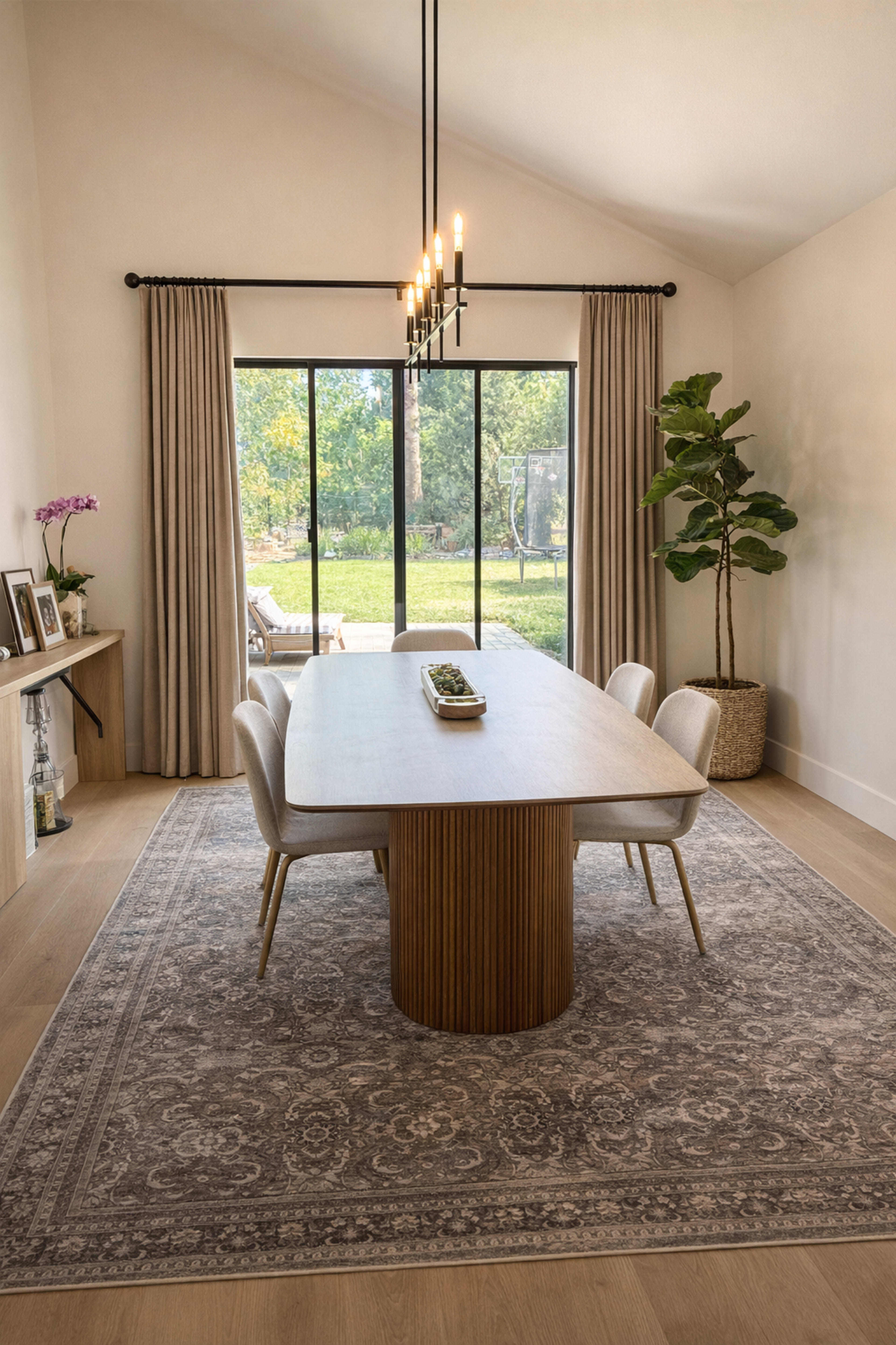 A modern dining room features a rectangular table with a round pedestal base, surrounded by four chairs, with large windows overlooking a garden.