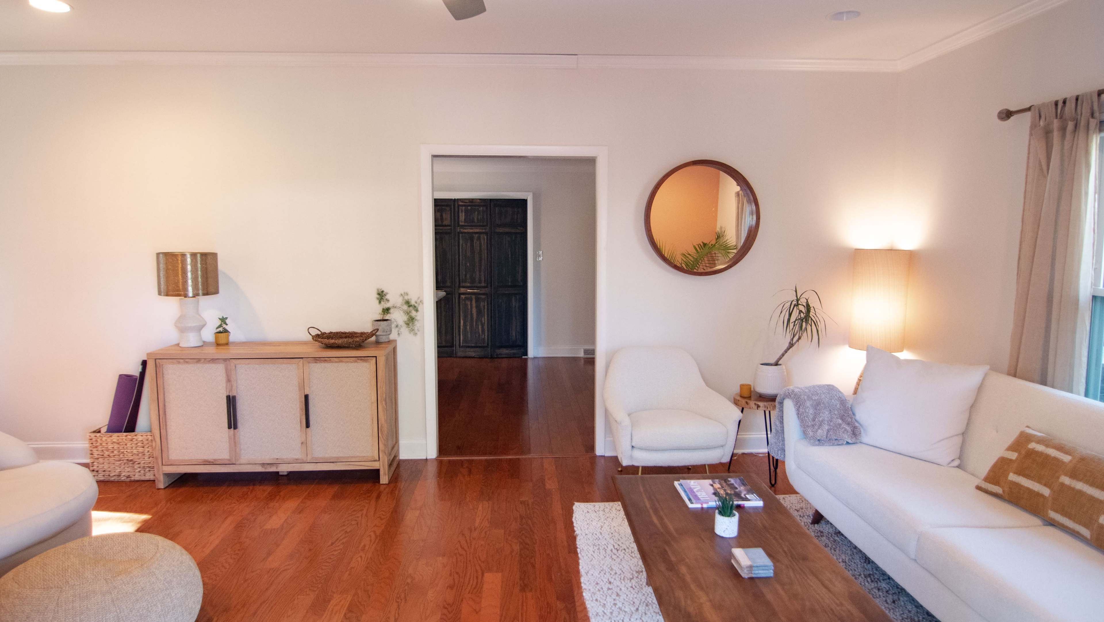 A living room with a light-colored sofa, wooden coffee table, a cabinet, and a round mirror on the wall.