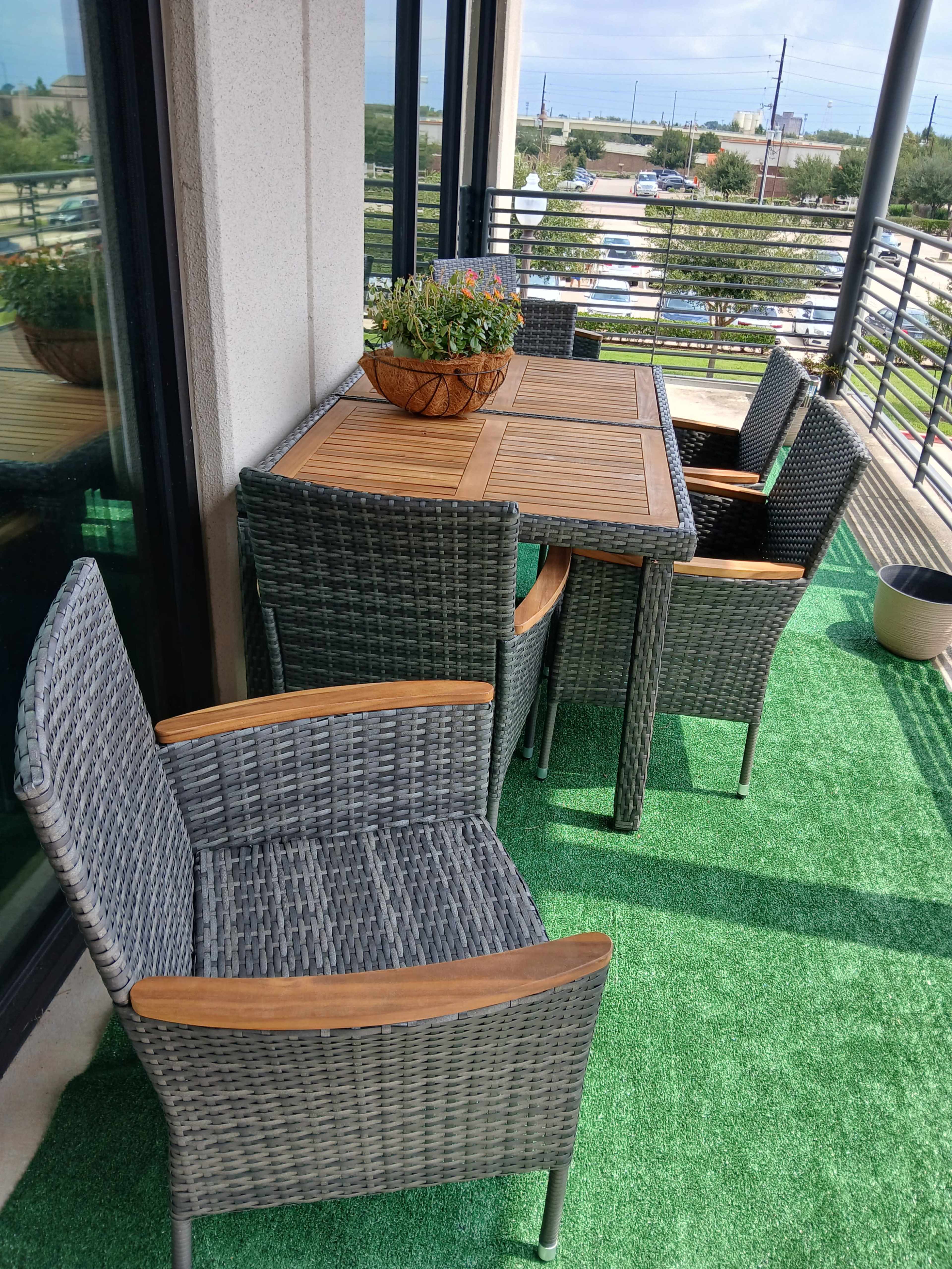 A balcony features a wooden table surrounded by four wicker chairs, with potted plants on the table and artificial grass covering the floor.