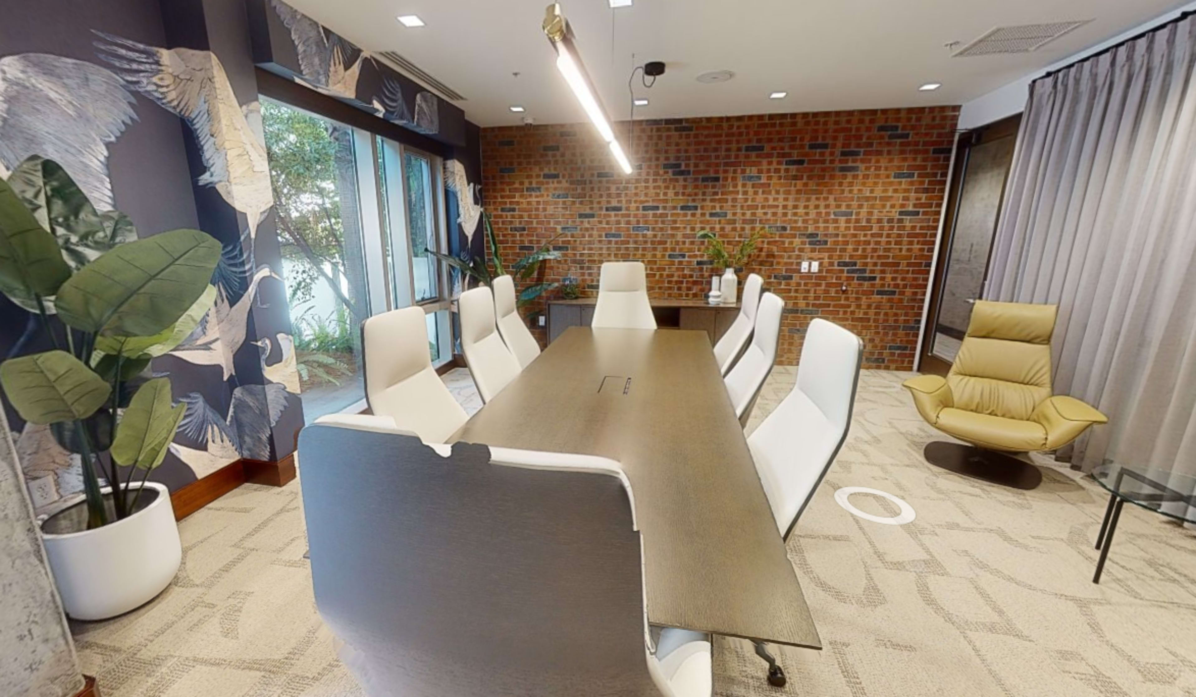 A modern conference room features a long table surrounded by white chairs, with a brick wall and plants in the background.