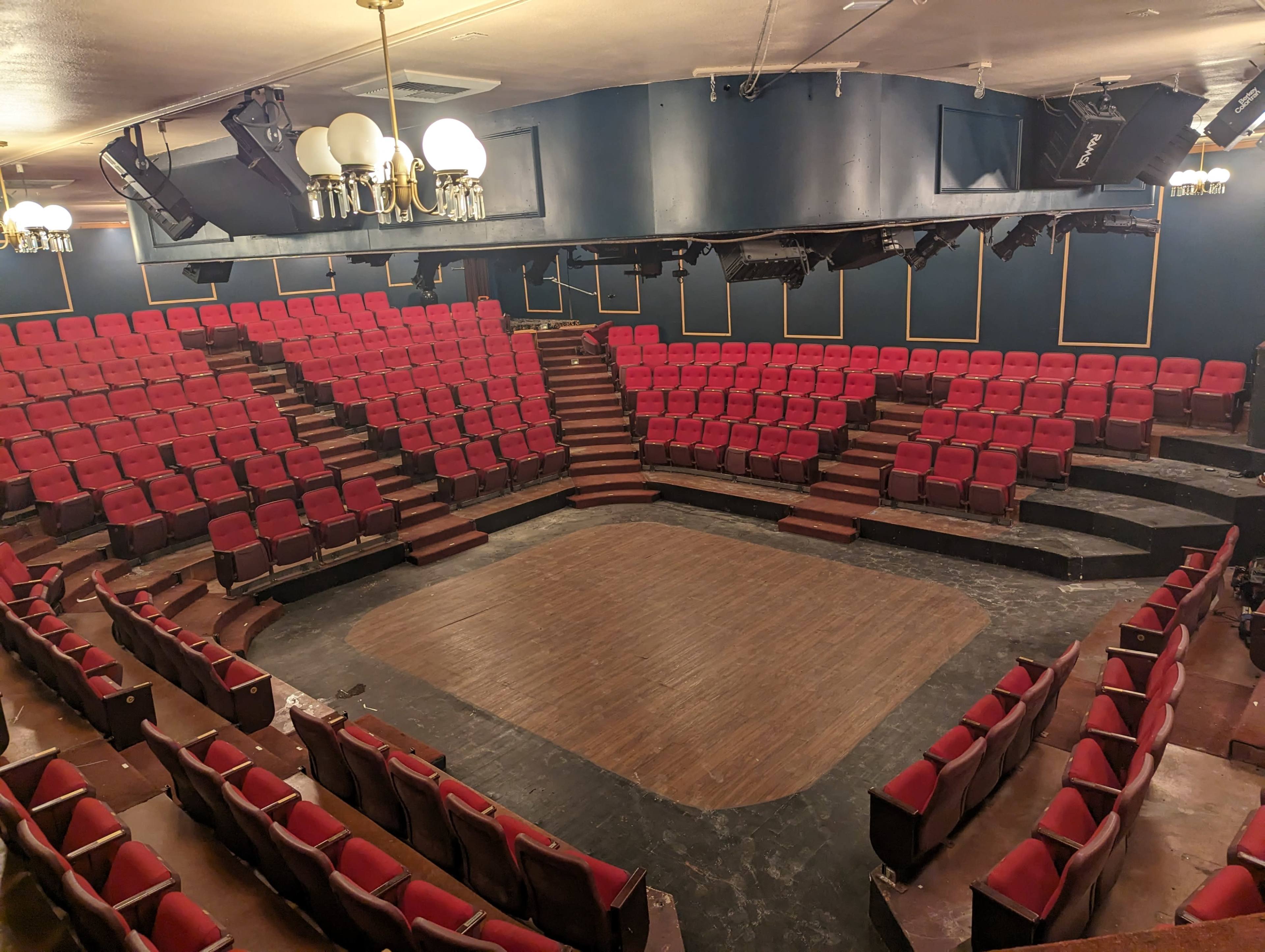 The image shows a theater with a circular seating arrangement, featuring red upholstered chairs and a wooden stage at the center.