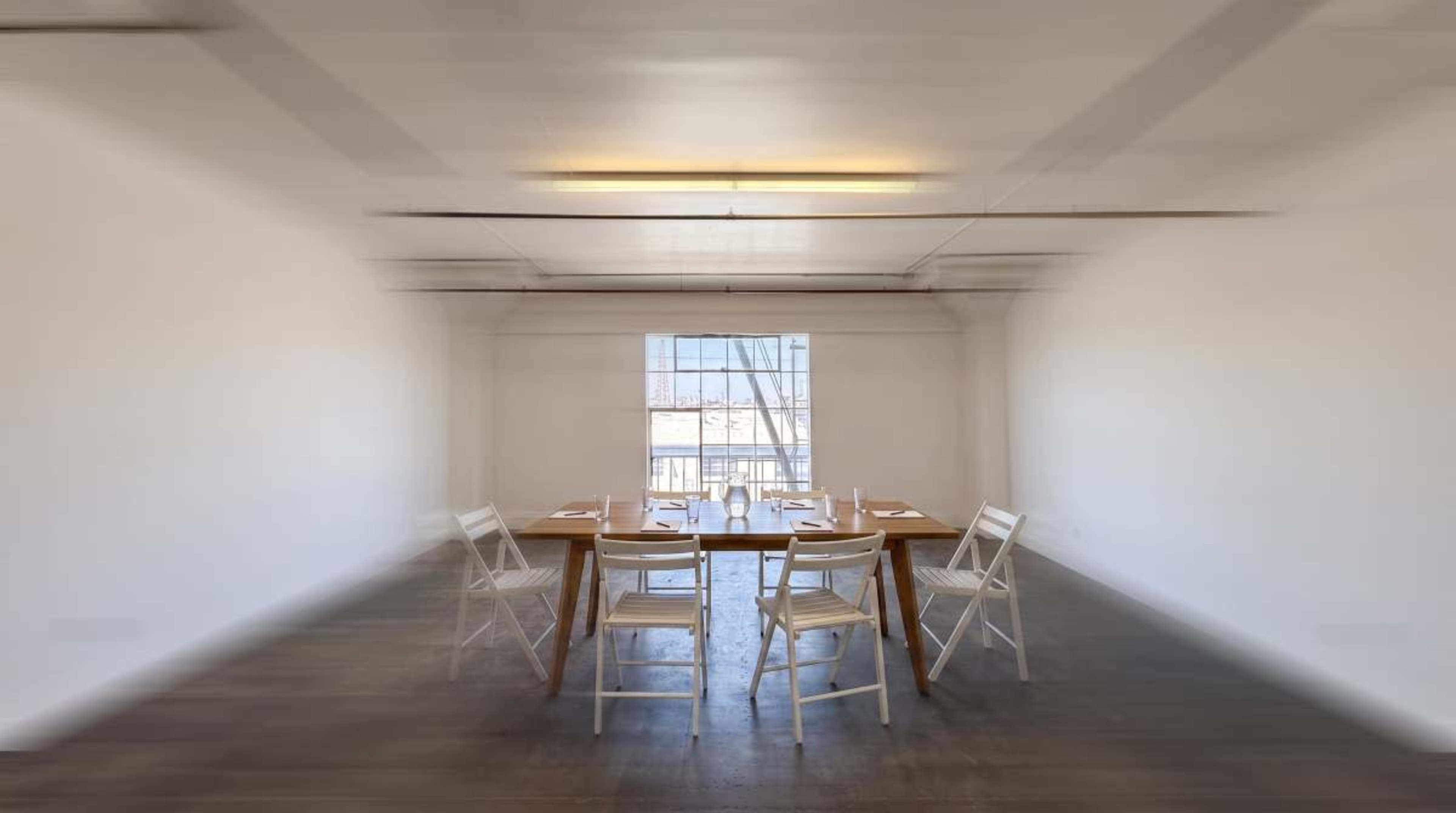 A wooden table is set with utensils and surrounded by white chairs in a bright, minimalist room with large windows.