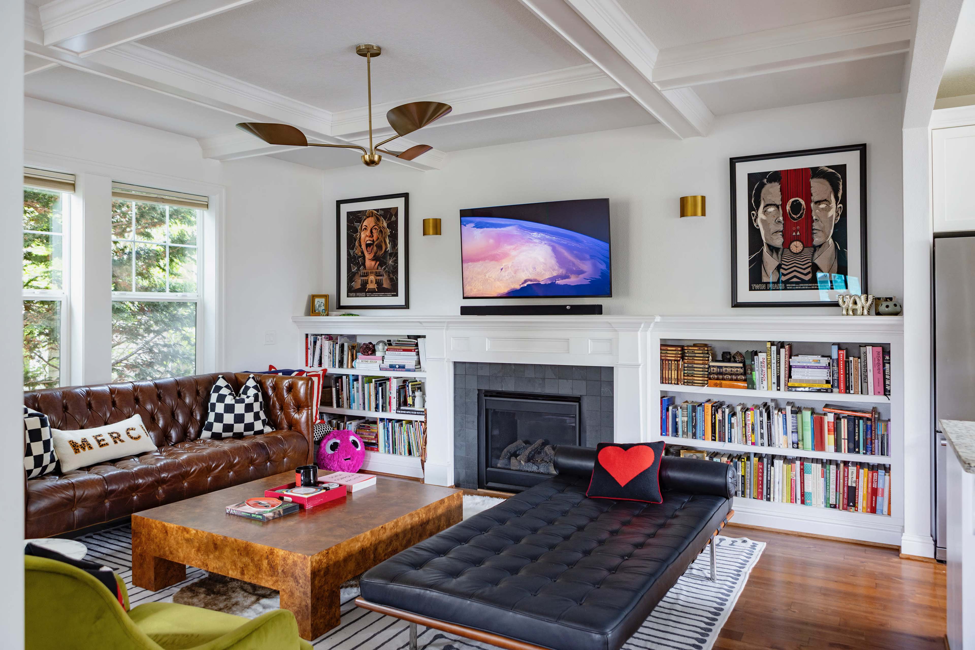 The image shows a modern living room featuring a brown leather sofa, a dark coffee table, a flat-screen TV mounted above a white fireplace, and bookshelves filled with books and decorative items.