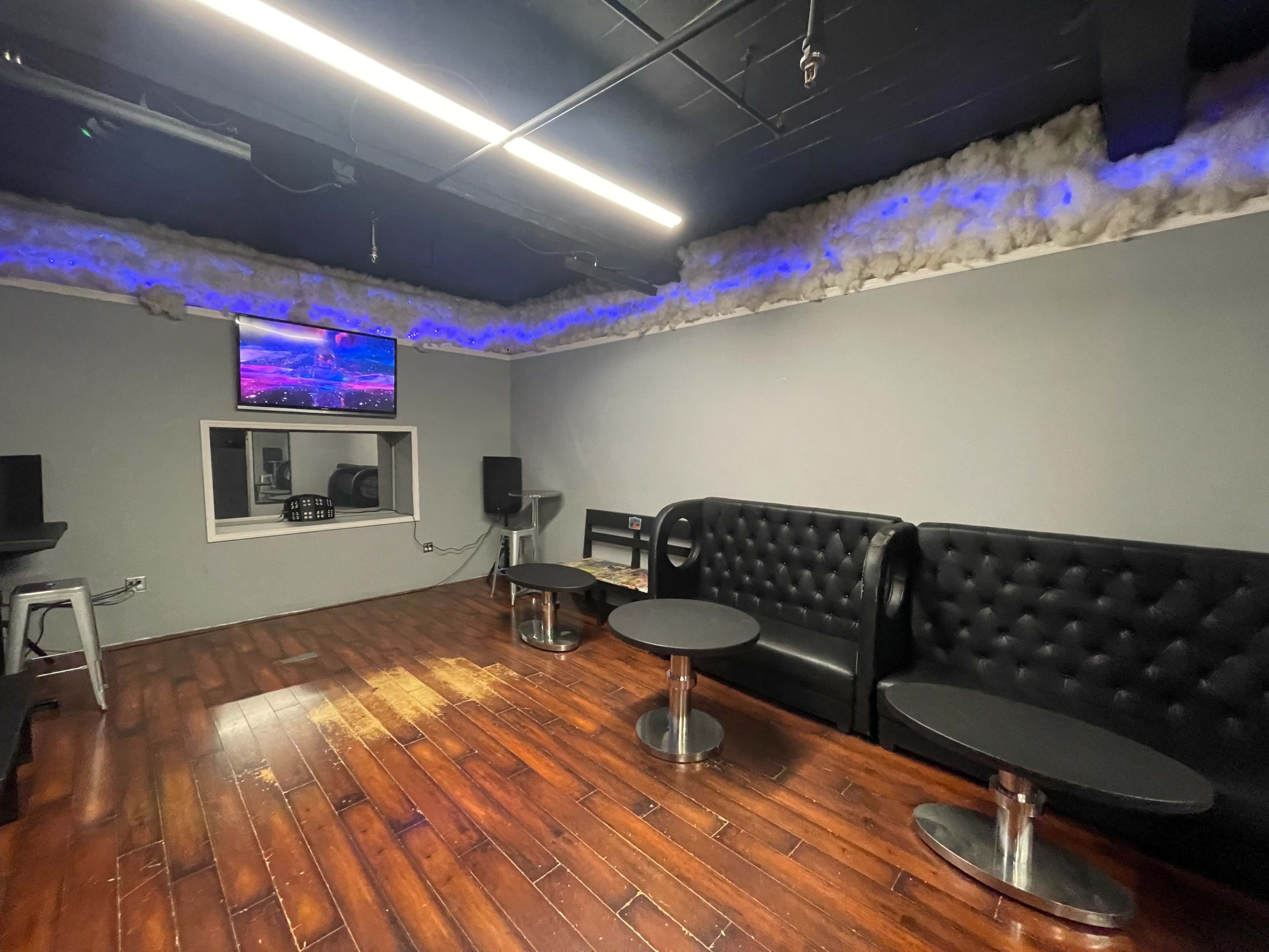 A modern lounge area with black tufted couches, two small tables, and a television mounted on the wall, illuminated by ambient lighting.