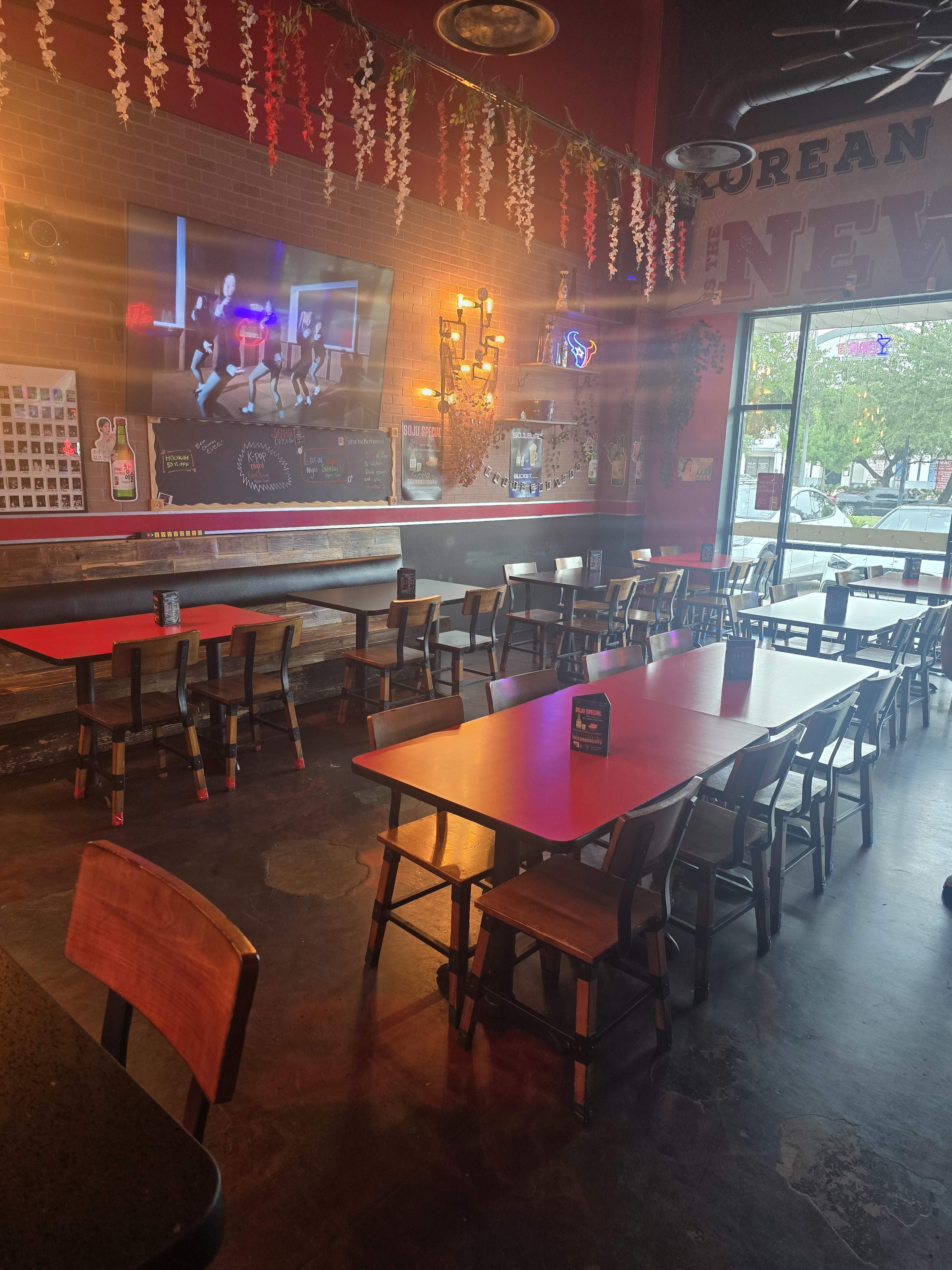 The image shows a restaurant interior featuring several tables and chairs, a large television on the wall, and decorative elements hanging from the ceiling.