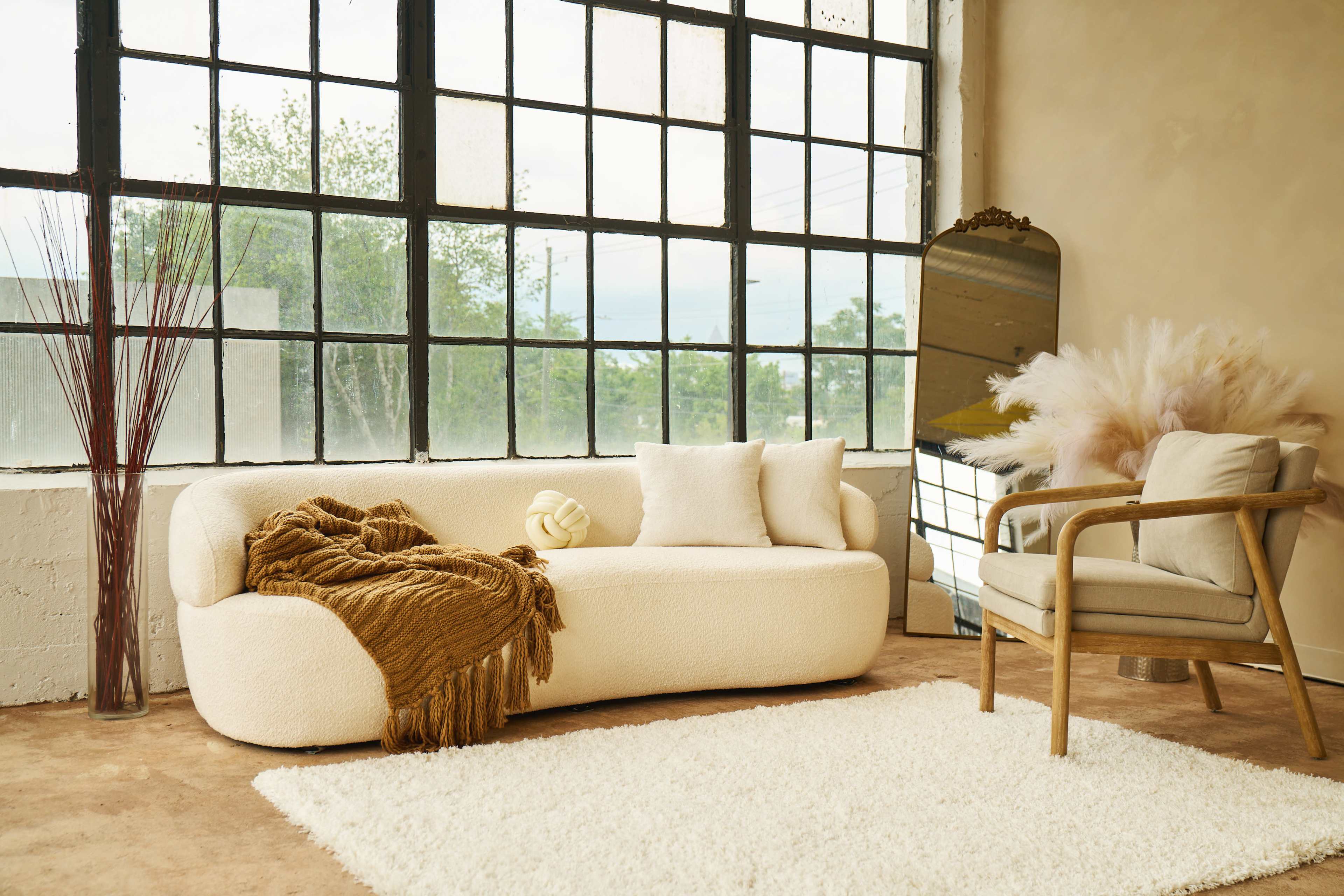 The image shows a cozy living area featuring a boucle-style sofa, a light-colored armchair, a large mirror, and decorative elements like a vase and a fluffy rug.