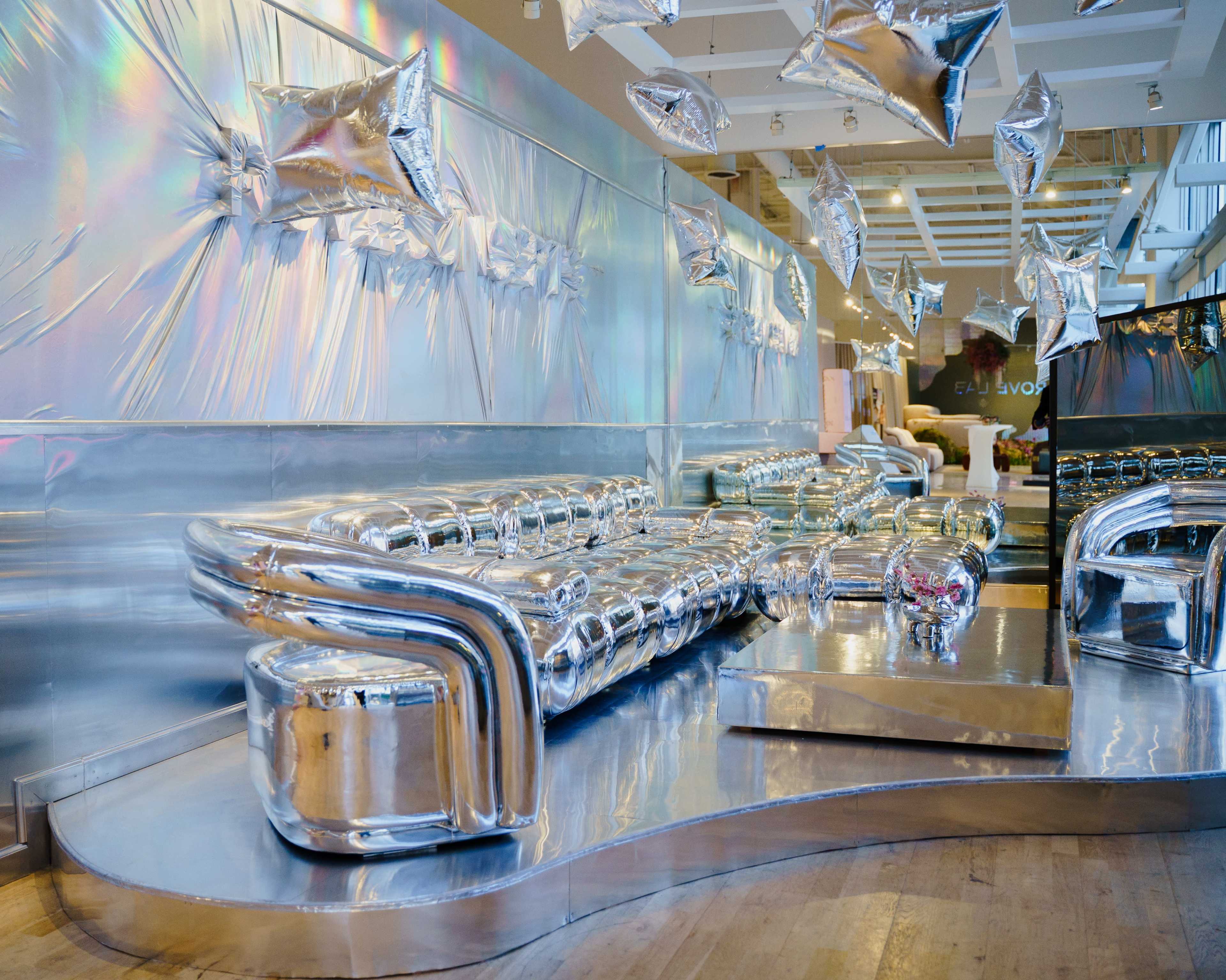 The image features a modern interior space with silver metallic furniture, including a sofa and decorative elements resembling stars, against a shiny reflective backdrop.