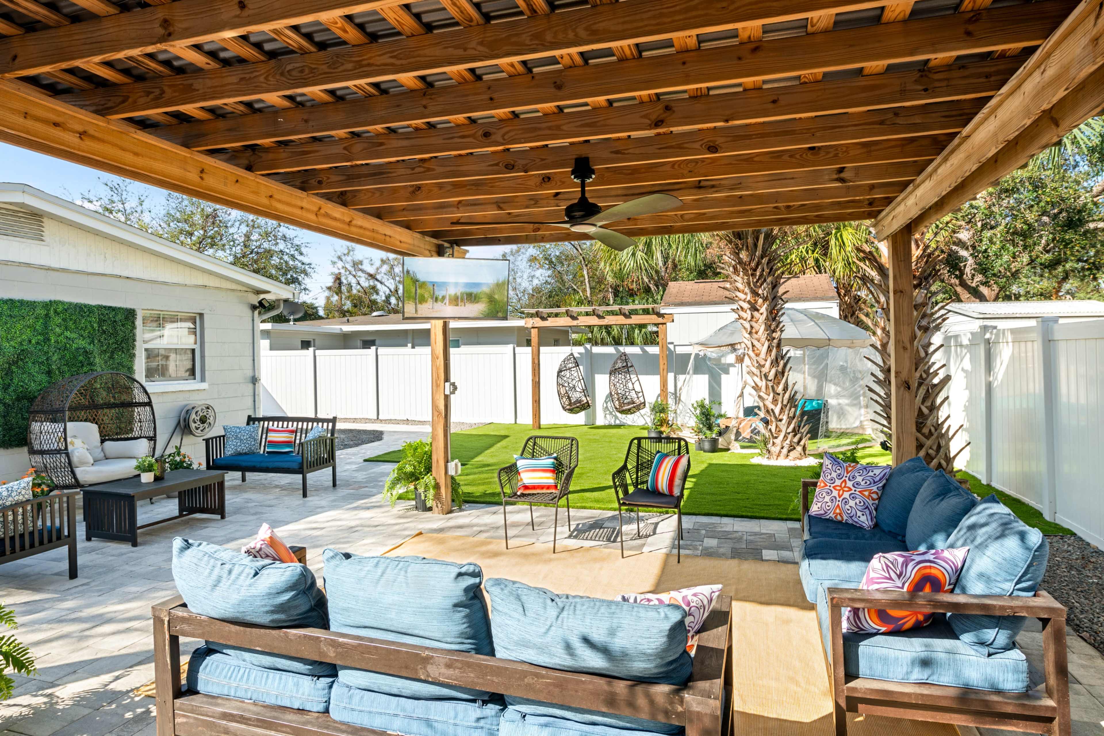 A covered outdoor seating area features a blue sofa, colorful cushions, and hanging chairs, surrounded by a grassy yard and palm trees.