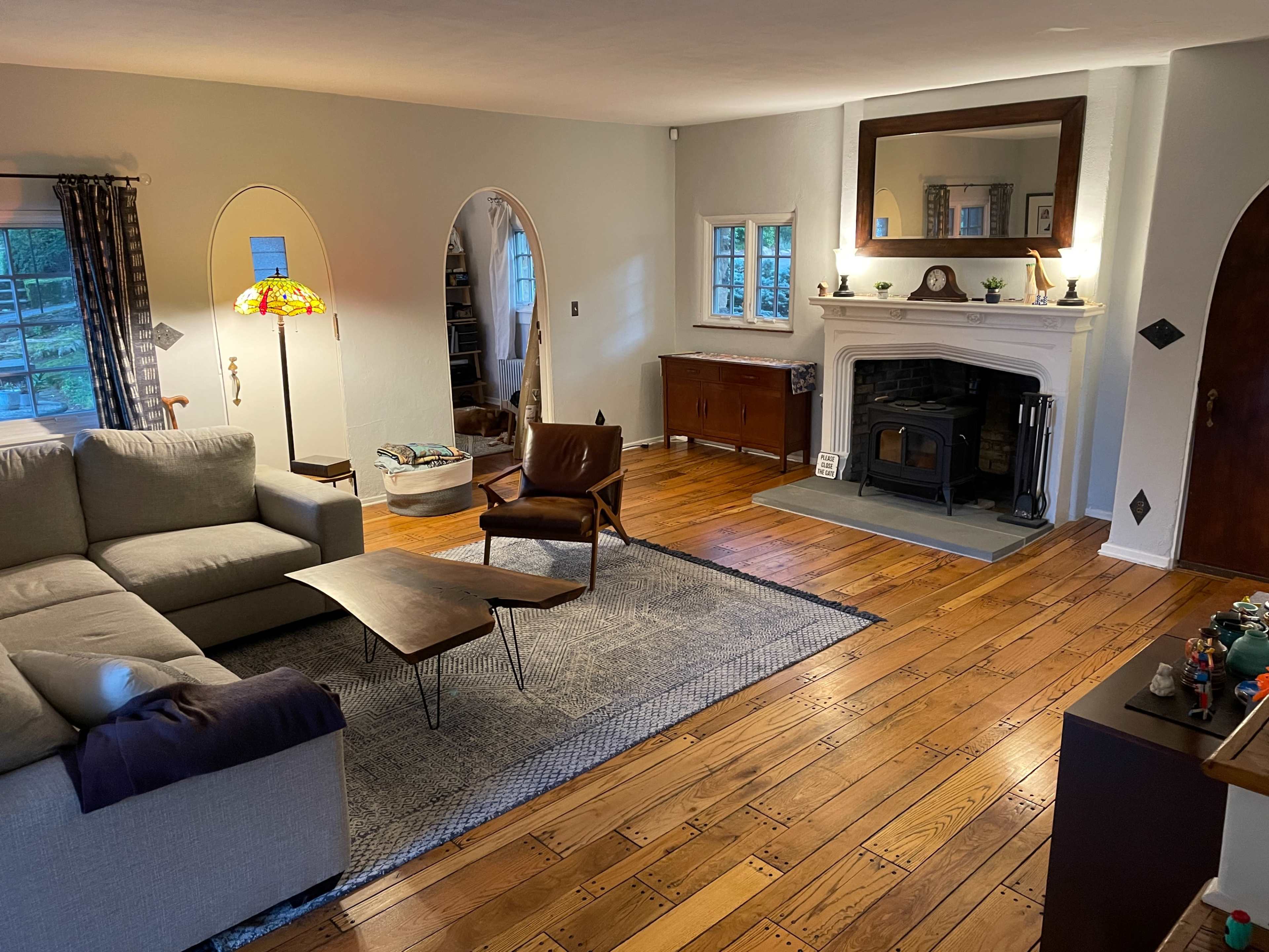 The image shows a cozy living room with a sofa, a coffee table, and a fireplace, complemented by wooden floors and a large mirror above the mantel.