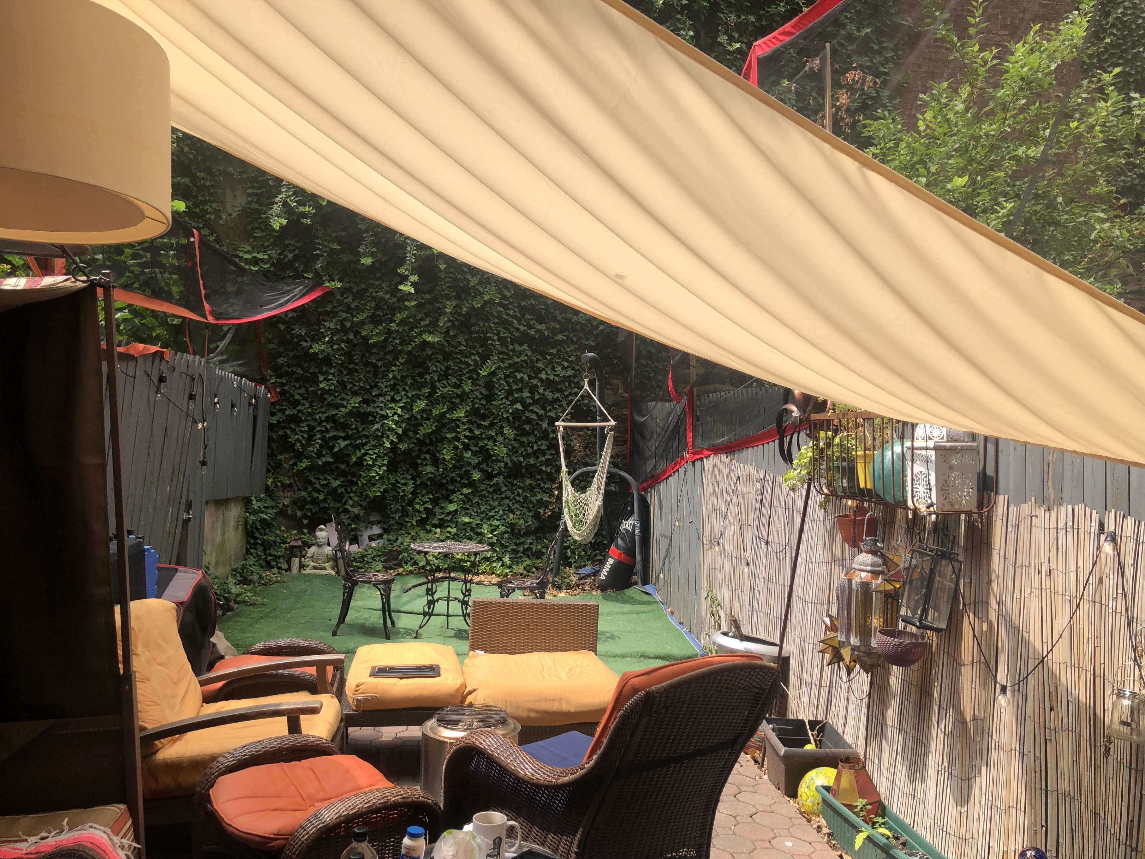 A small outdoor patio area with a hanging chair, several seating options, and a canopy overhead, surrounded by greenery and decorative elements along the walls.