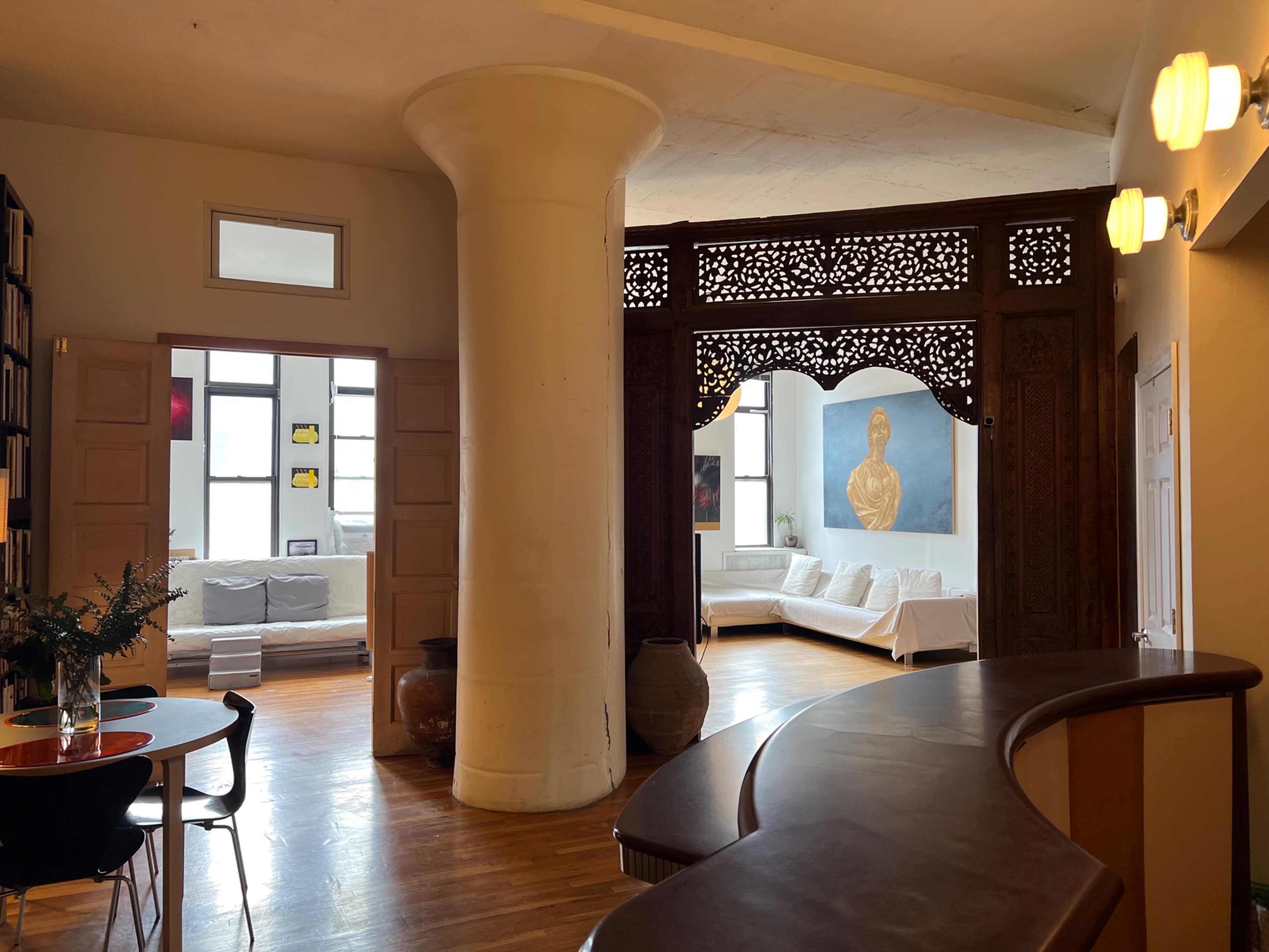 The image shows a spacious room with a large ornate wooden divider, a circular column, and a seating area featuring a white couch and a large painting of a figure.