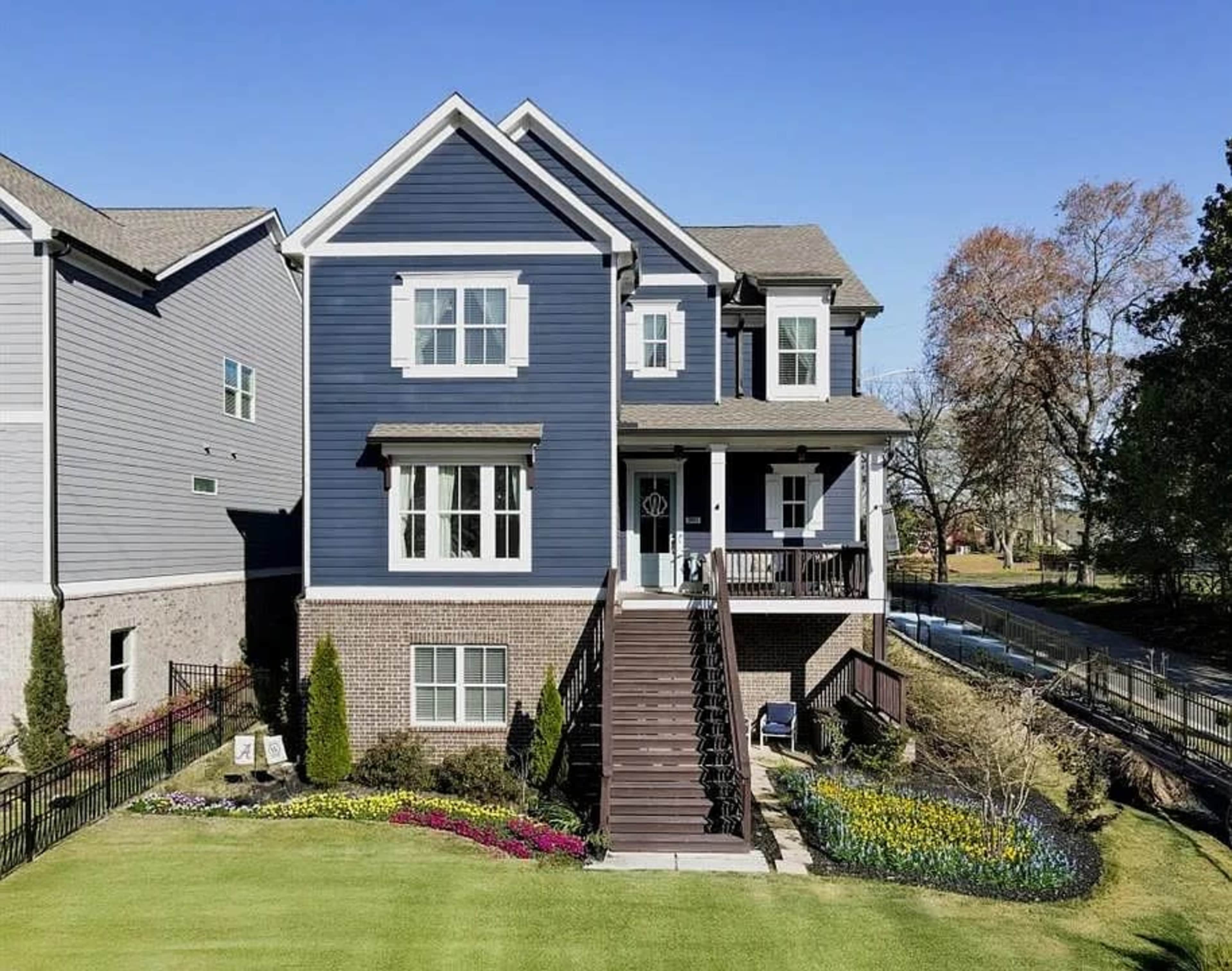 A two-story blue house with a porch and large windows is surrounded by a neatly landscaped yard featuring flowers and shrubs.
