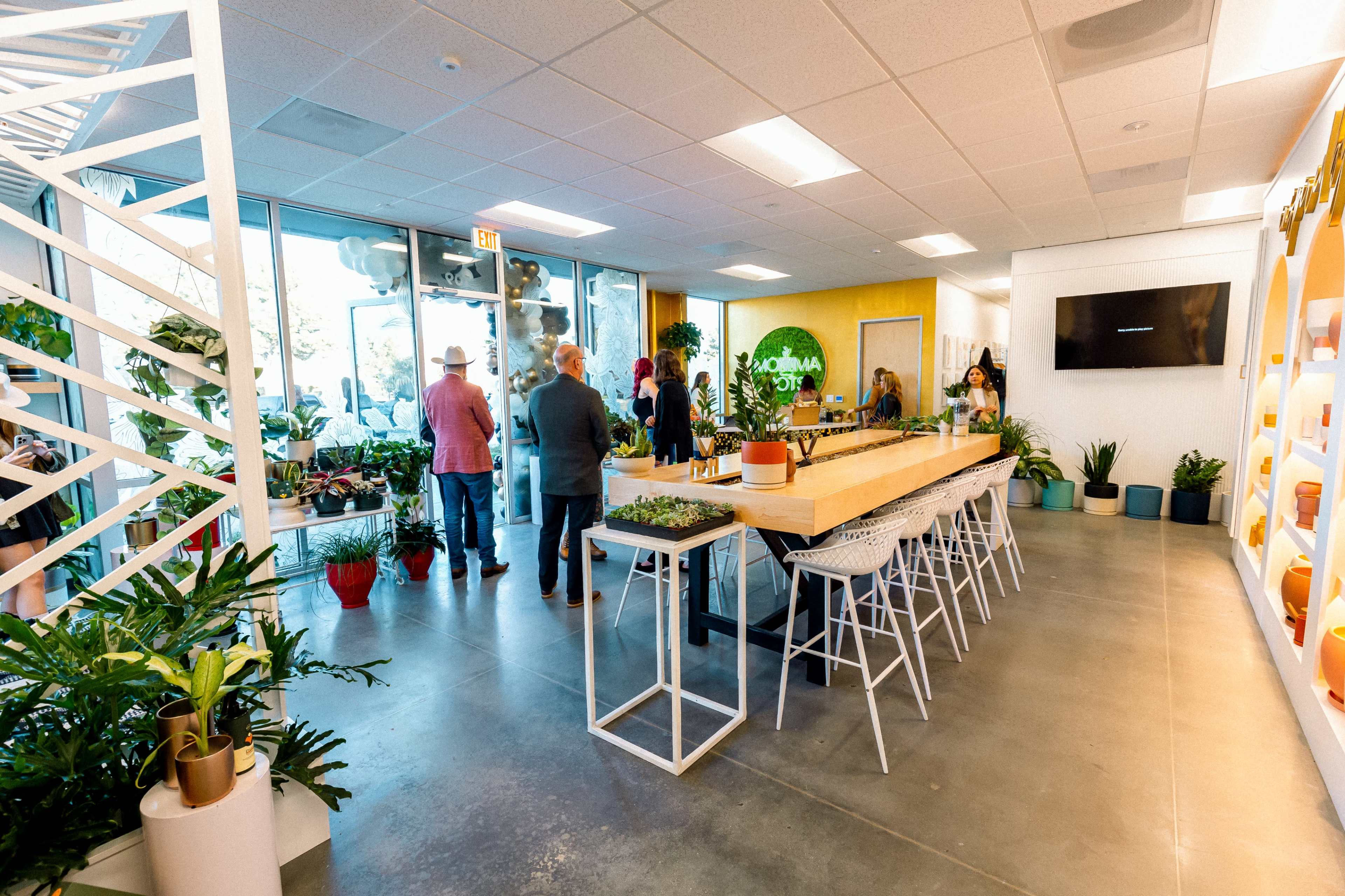 A group of people gathers in a modern, bright indoor space adorned with plants and a long wooden table surrounded by white stools.