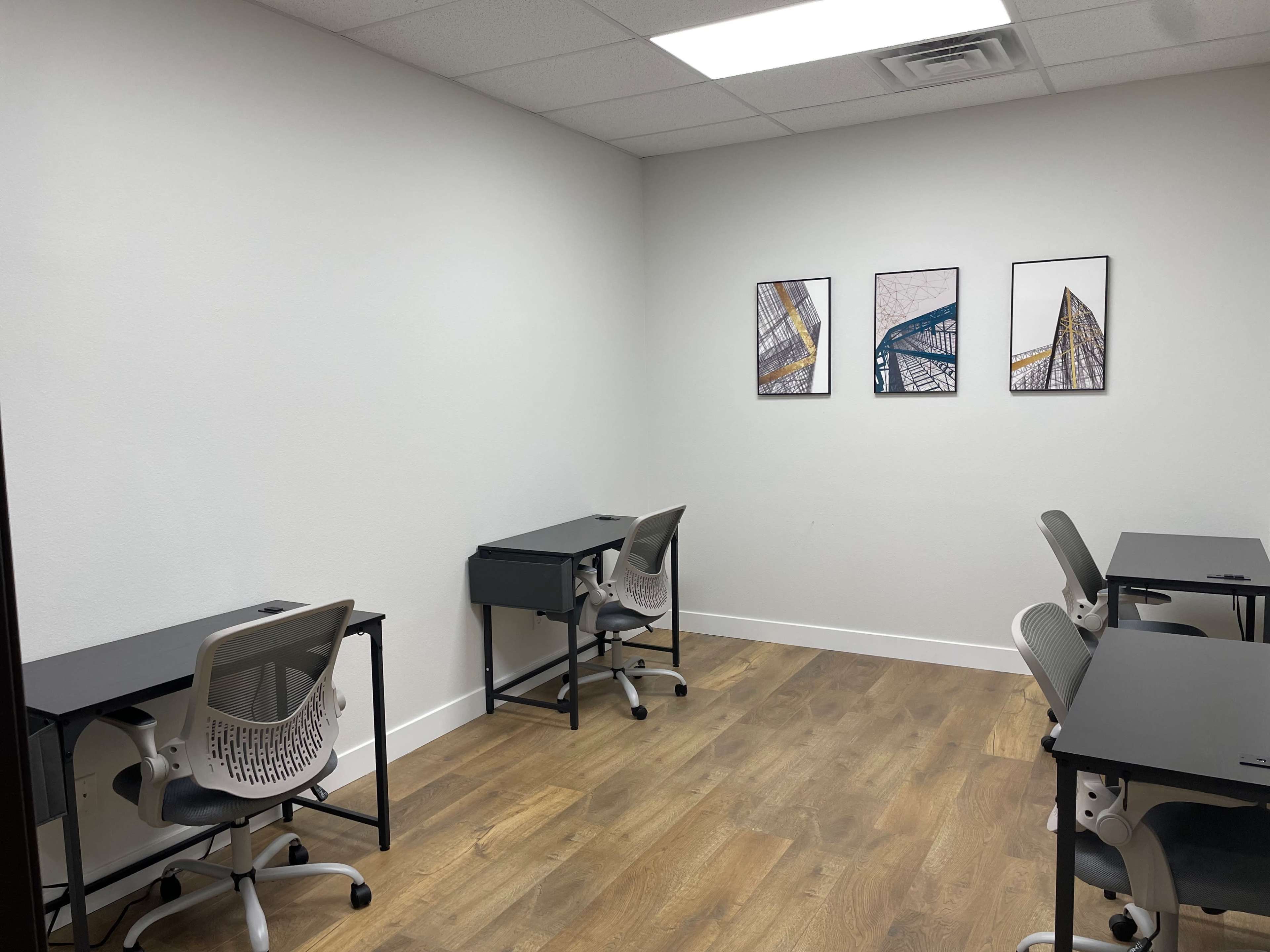 A minimalistic office space with four black desks and rolling chairs arranged against white walls, featuring three framed pictures on one wall.