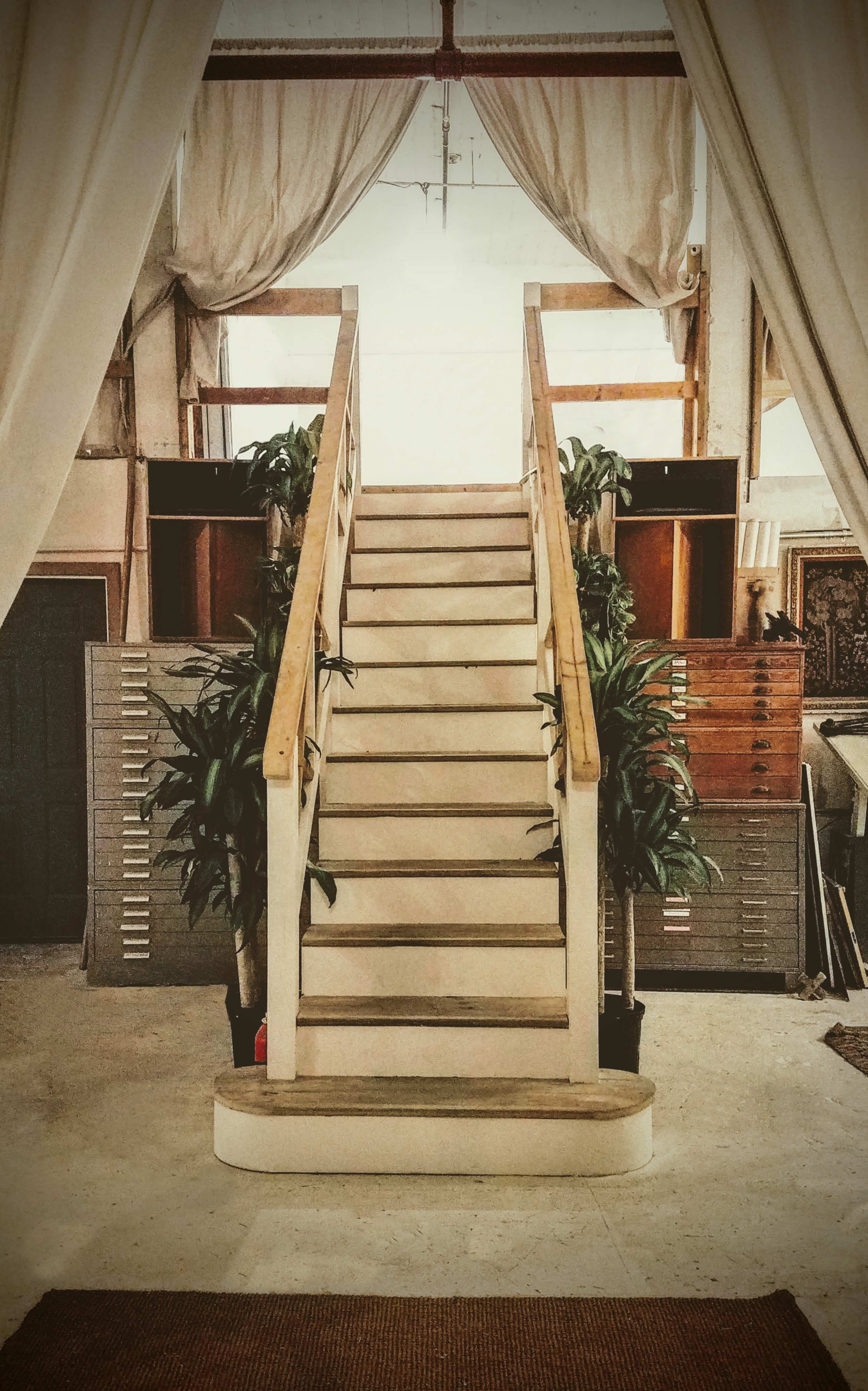 A staircase with a wooden railing, flanked by potted plants, leading to an upper level in a bright, spacious room.