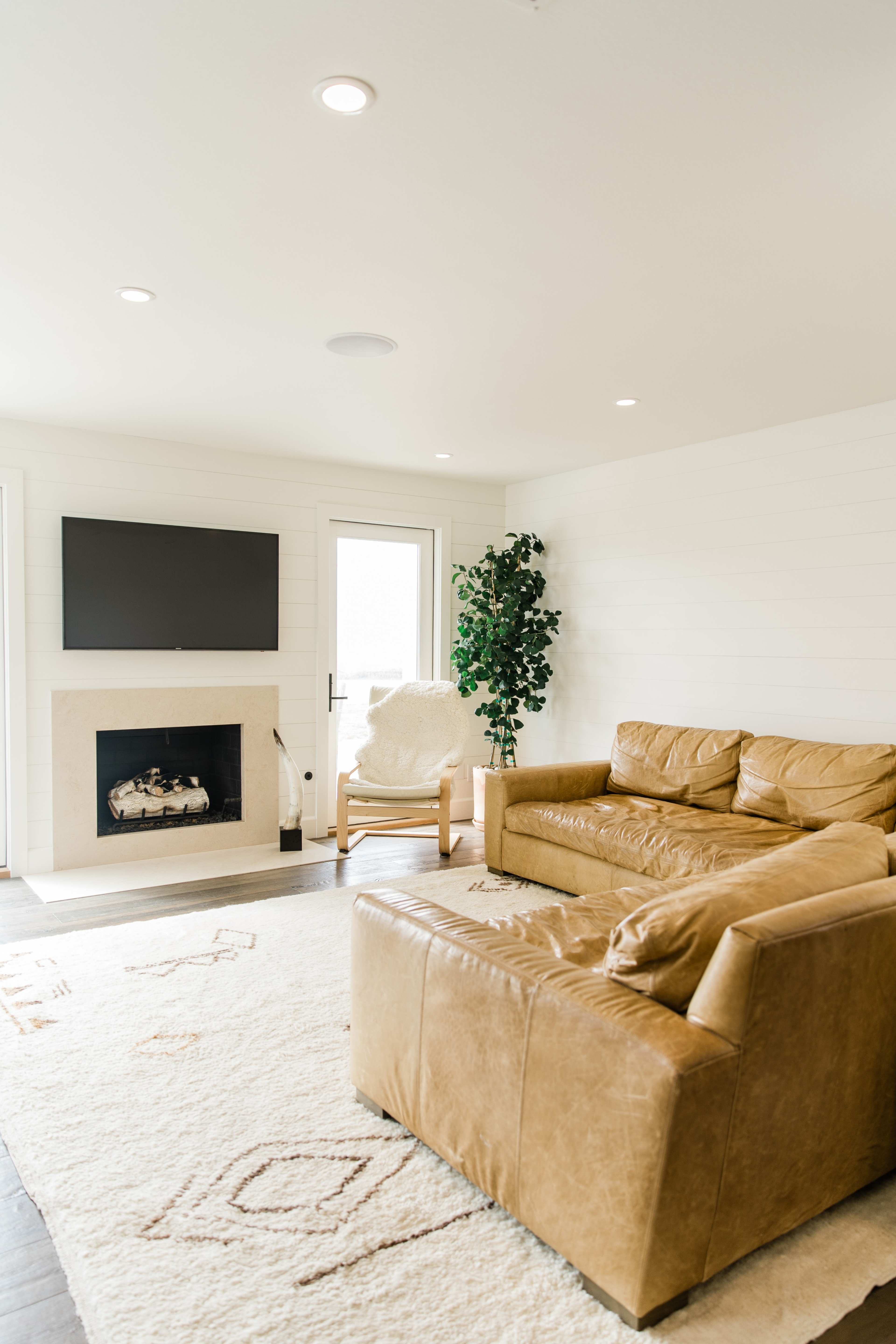 A light-filled living room with a beige sectional sofa, a flat-screen TV mounted on the wall, and a cozy armchair beside a decorative plant.