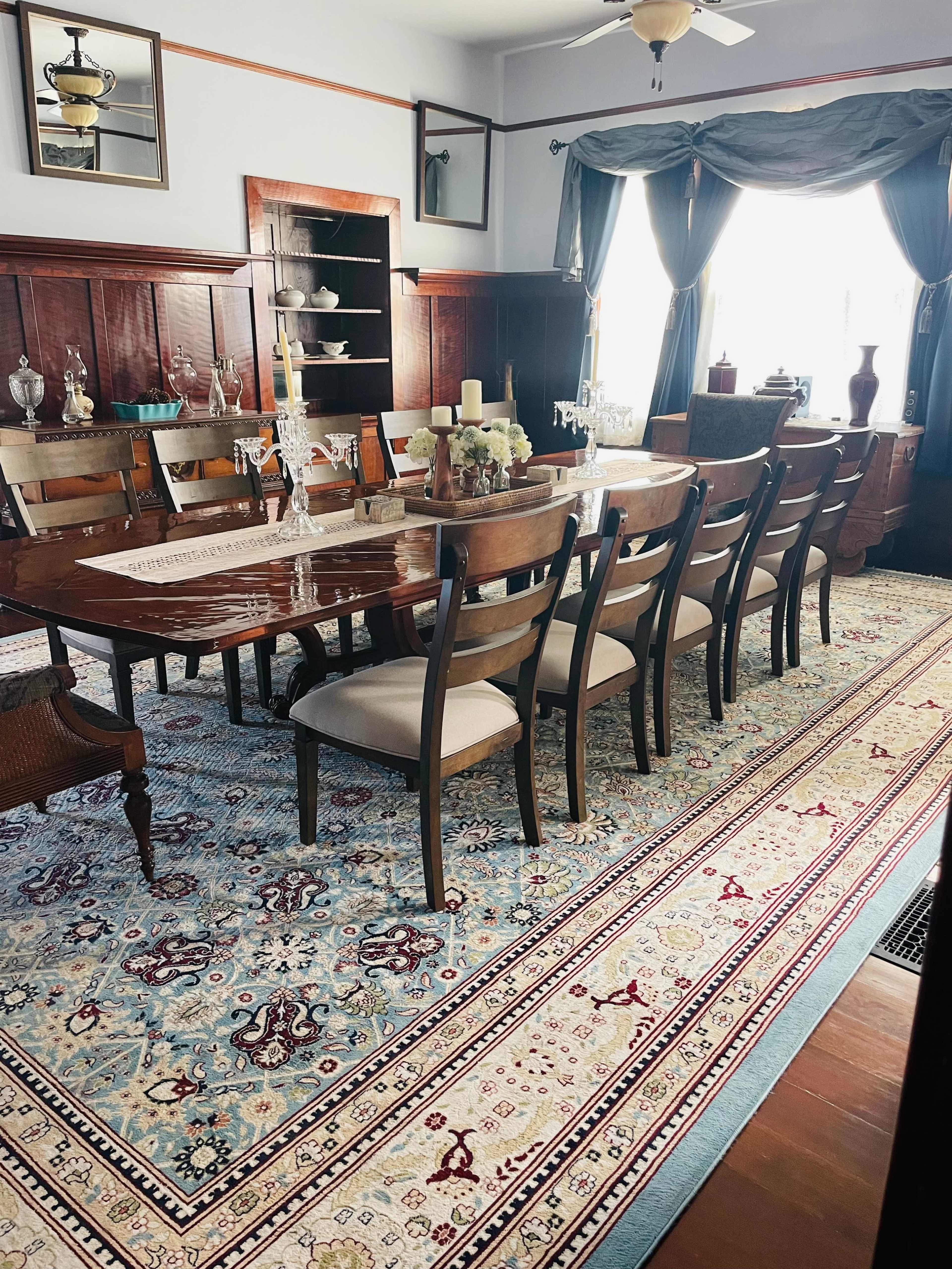 A large dining room features a polished wooden table surrounded by ten chairs, a patterned rug, and elegant decor along the walls.