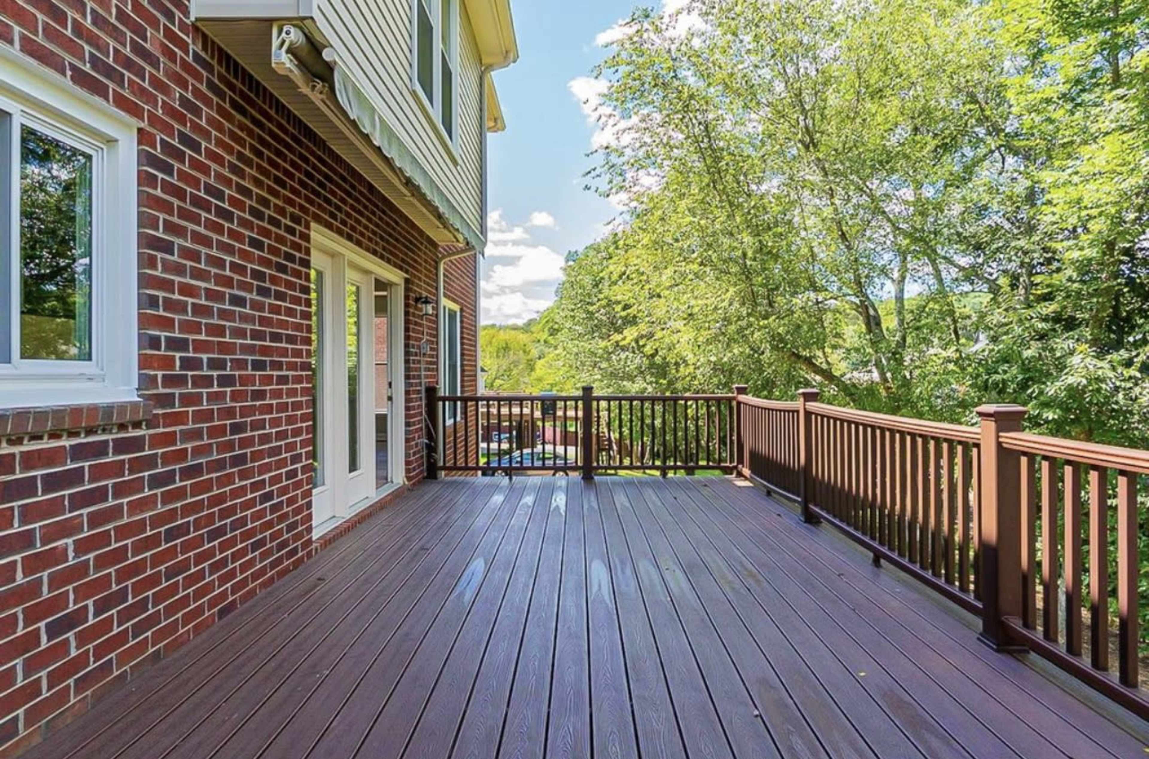 A wooden deck with a railing, adjacent to a brick house, surrounded by trees and greenery.