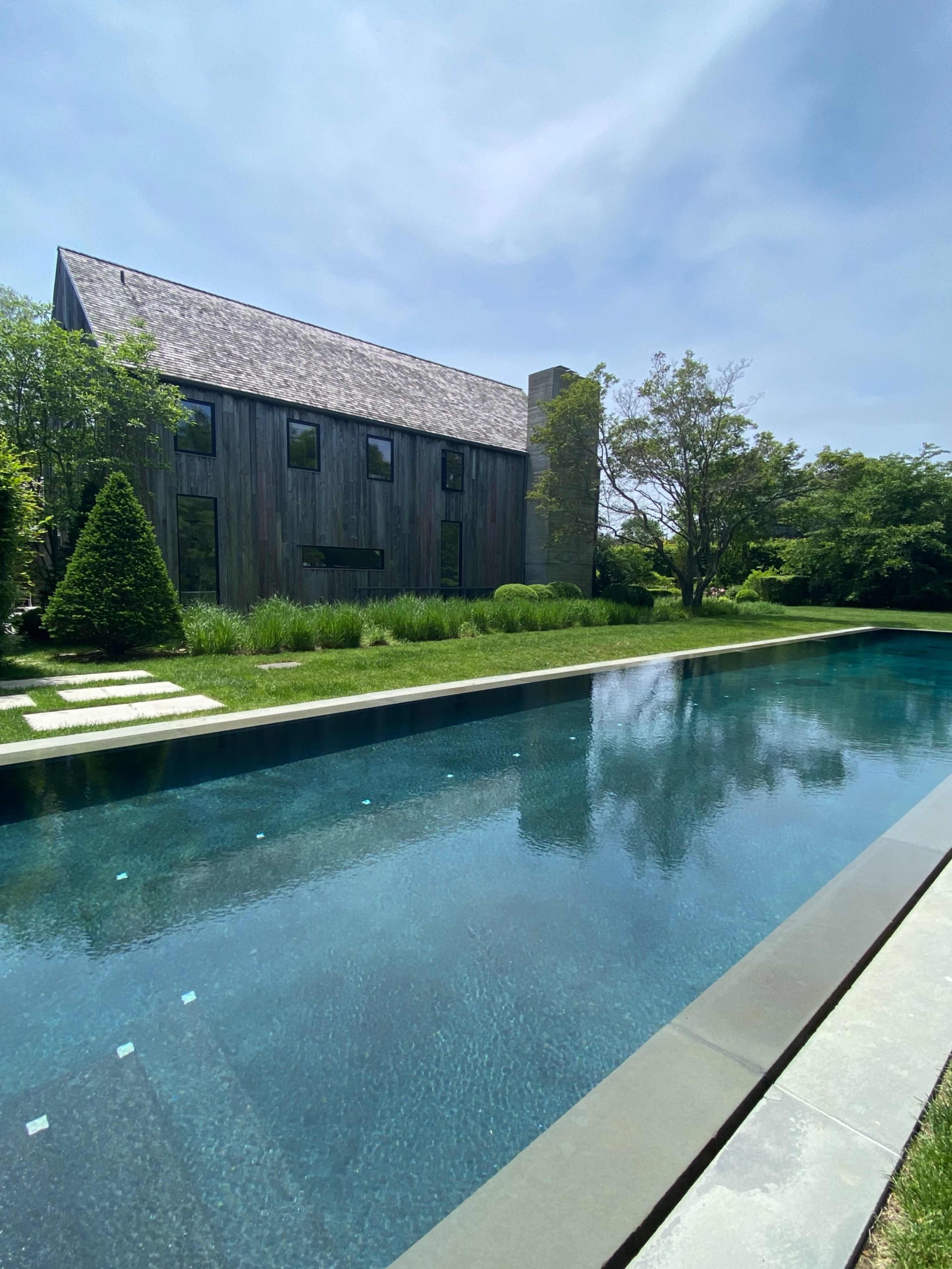 A modern wooden house stands beside a long, clear swimming pool surrounded by green landscaping.