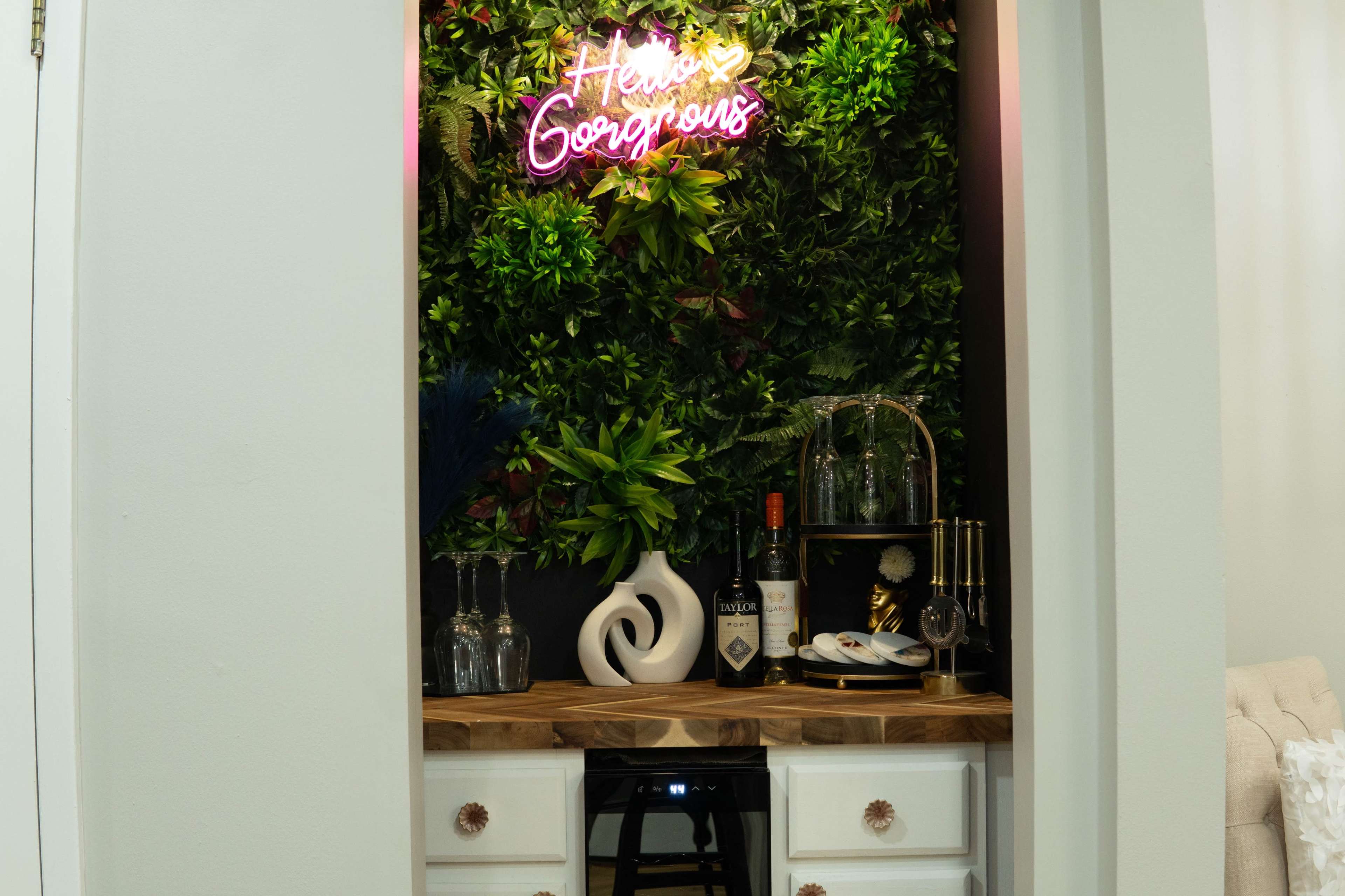 A small bar area with a wooden countertop, decorative bottles, and a neon sign that reads "Hello Gorgeous" is set against a backdrop of green plants.