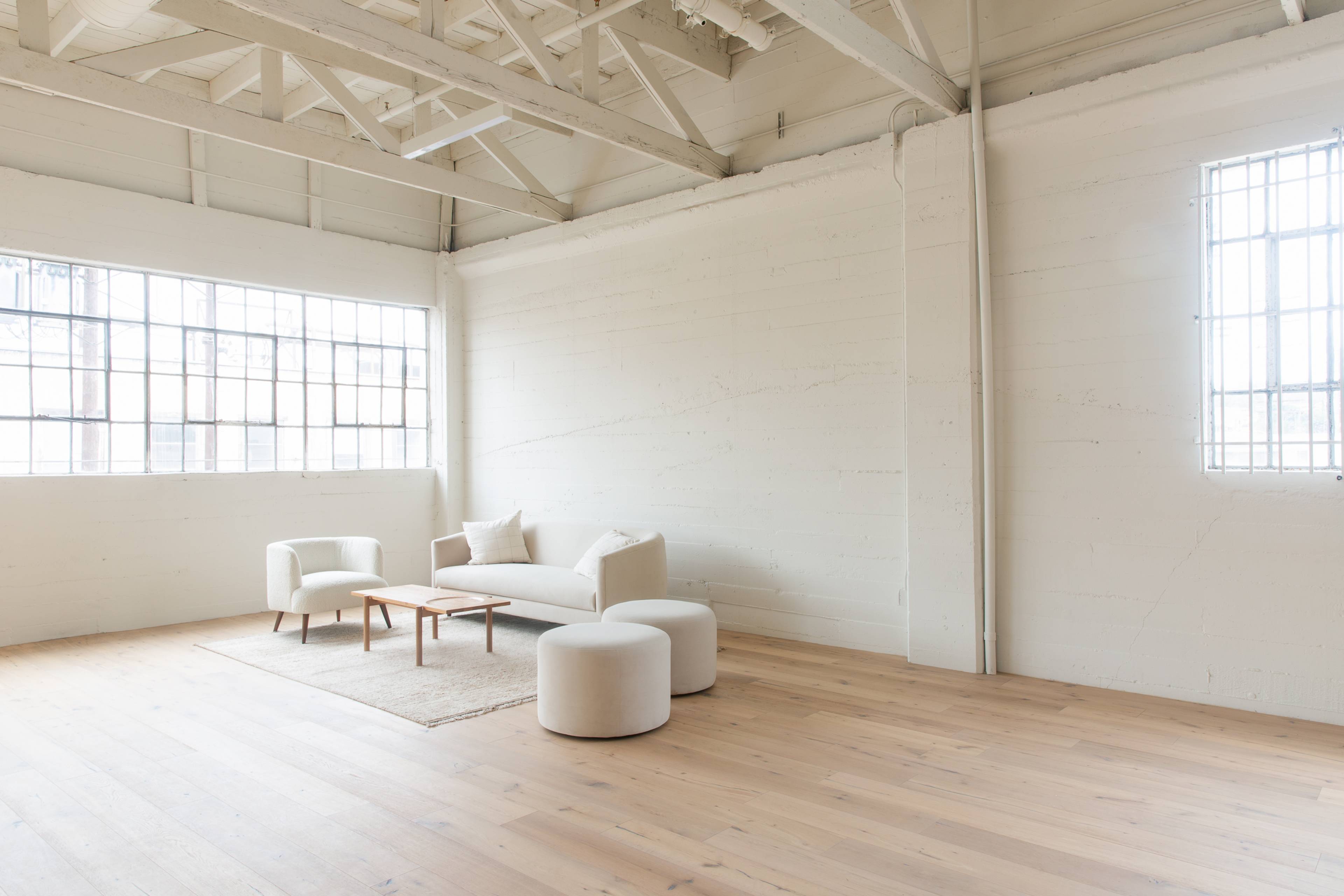 A bright, minimalist living space features a white sofa, two round ottomans, and a small wooden coffee table on a light wood floor.