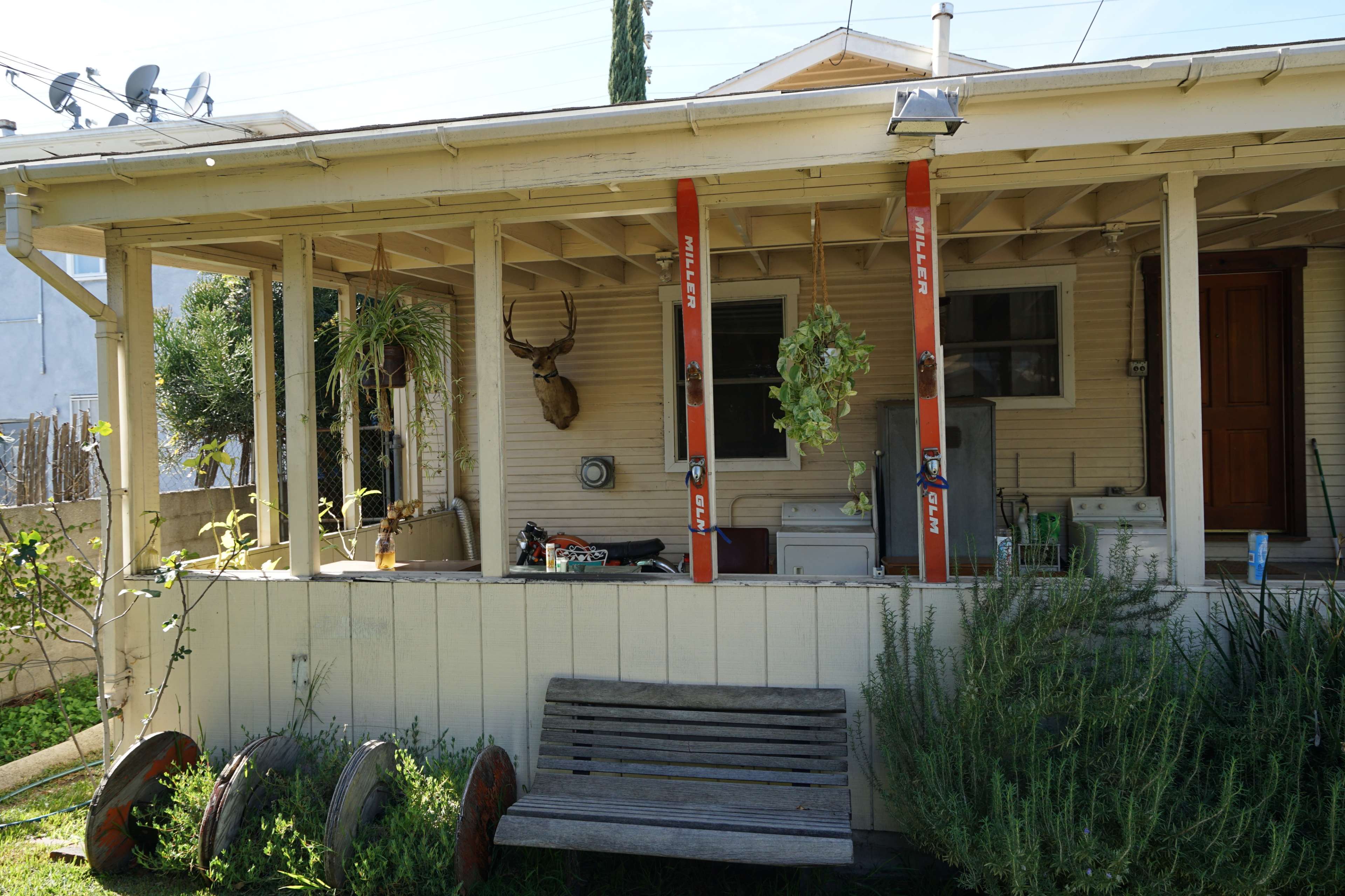 A porch with two vertical red posts, a mounted deer head, and a wooden bench is situated in front of a house surrounded by plants and equipment.