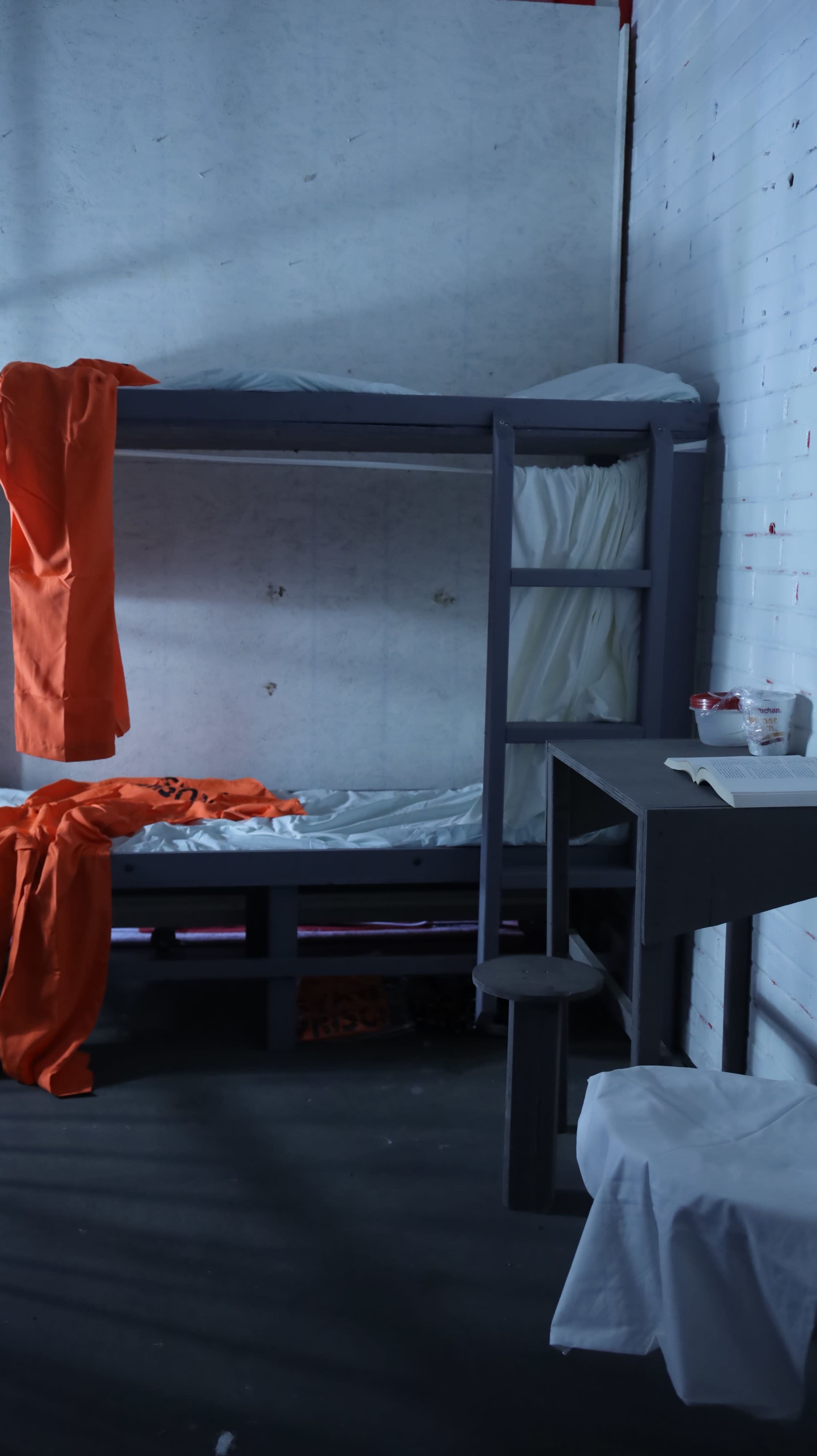 The image shows a small prison cell with two metal bunk beds, one covered with a white mattress, and orange prison uniforms hanging from the bed and a nearby table.