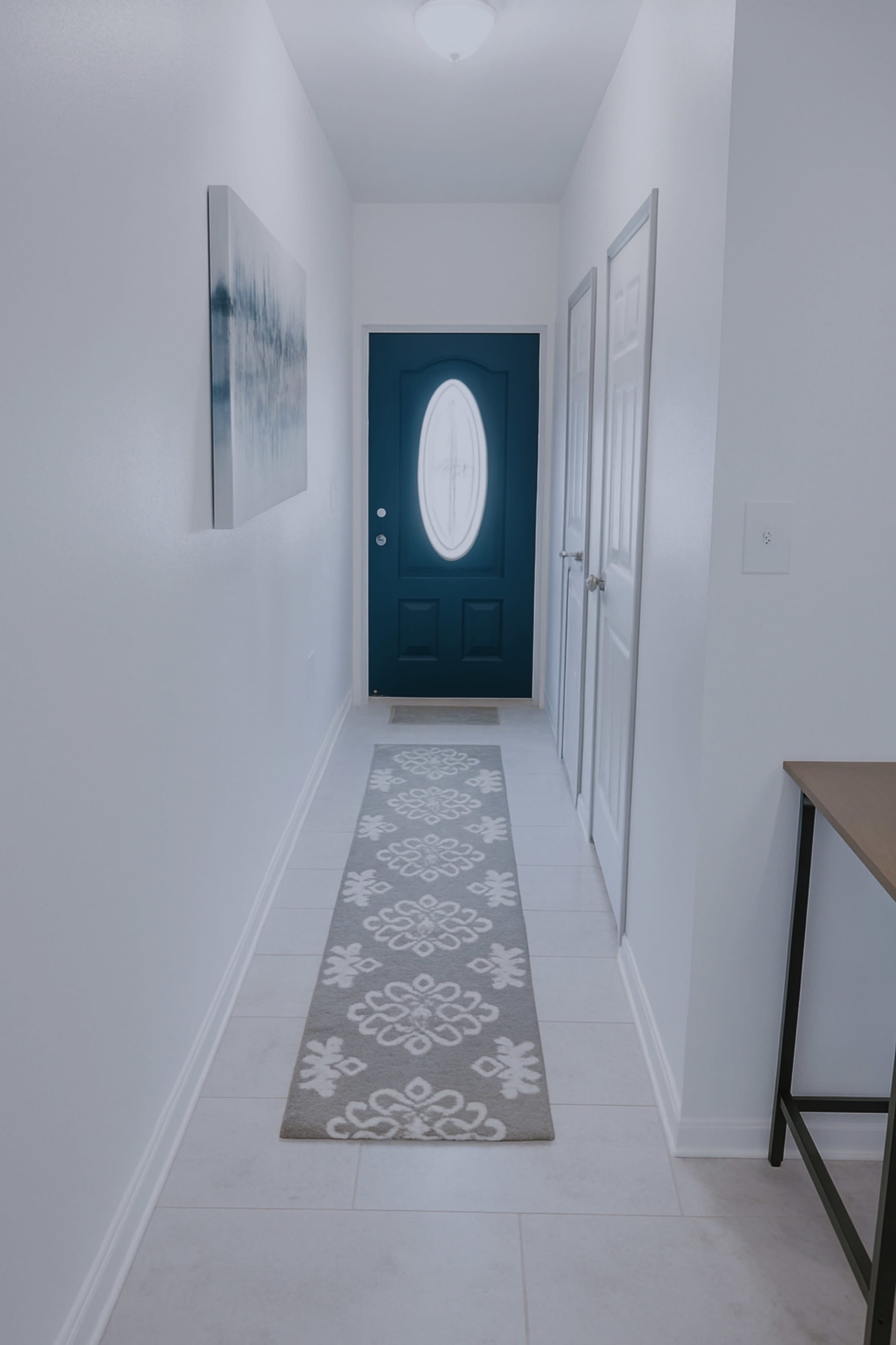 A long, narrow hallway features a blue front door at the end, flanked by two closed doors, with a light-colored rug laid along the tiled floor.