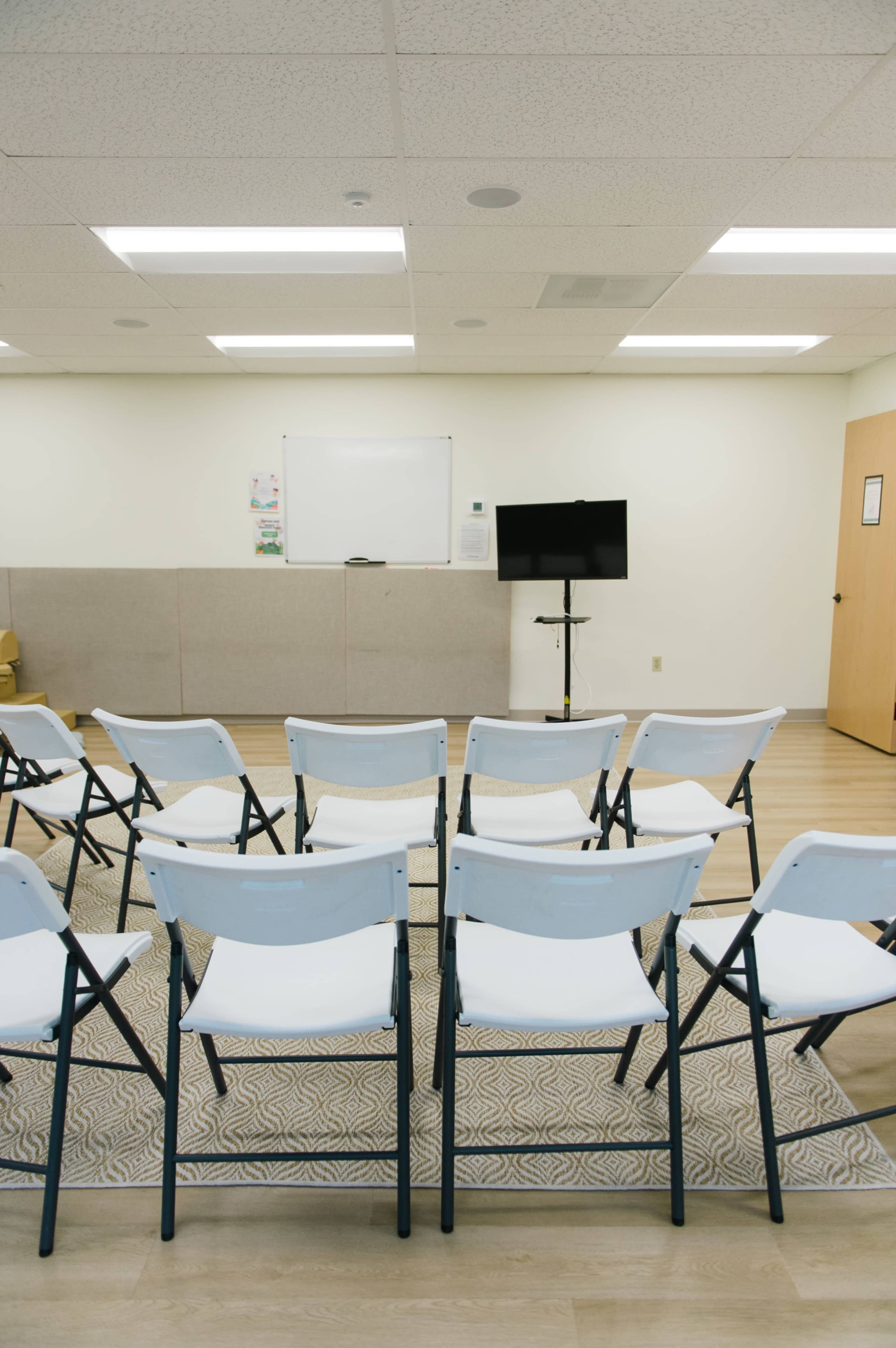 The image shows a row of empty folding chairs facing a blank whiteboard and a television on a stand in a brightly lit meeting room.