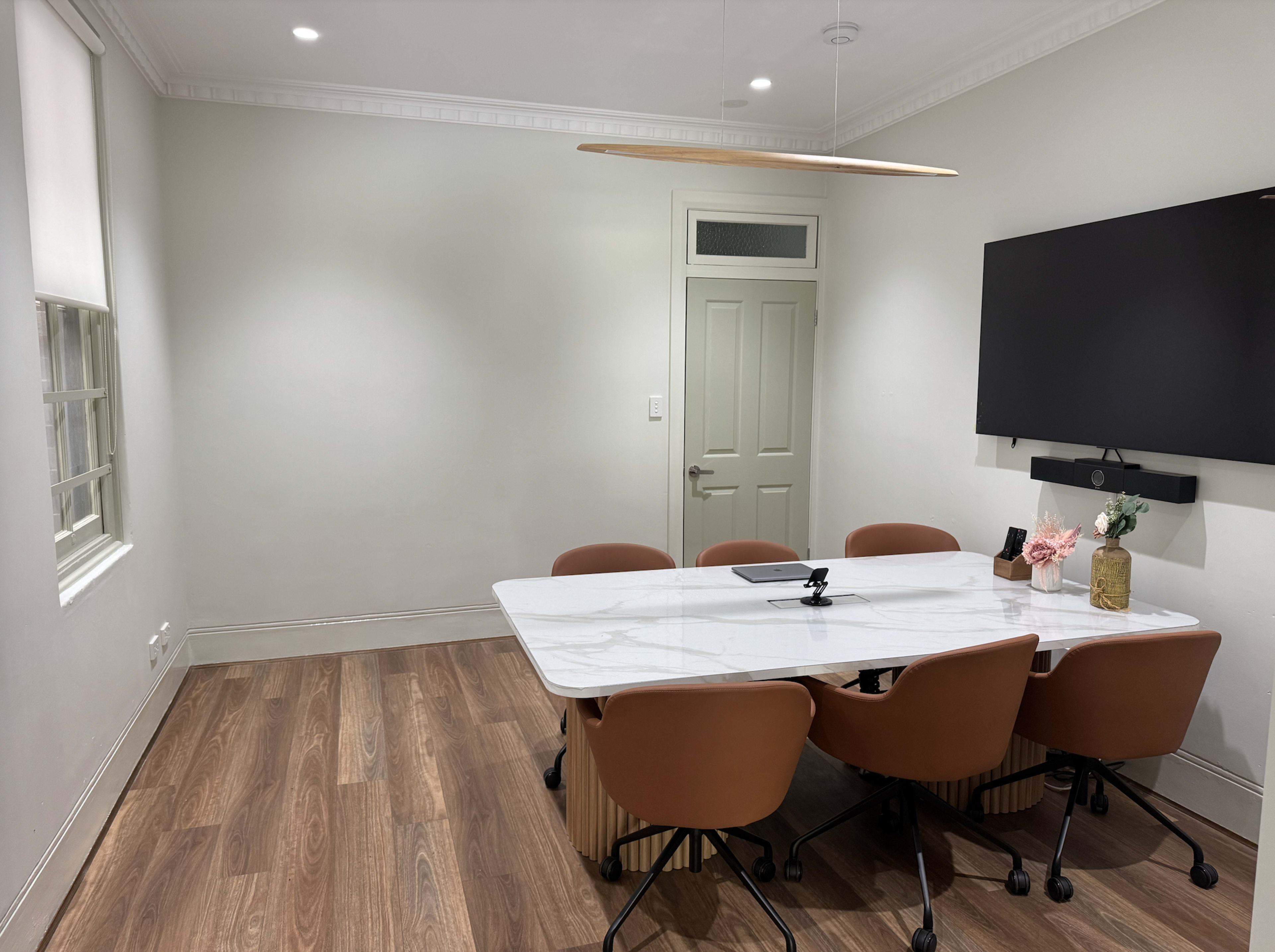 The image shows a modern conference room with a marble table, four brown chairs, a wall-mounted TV, and soft lighting.