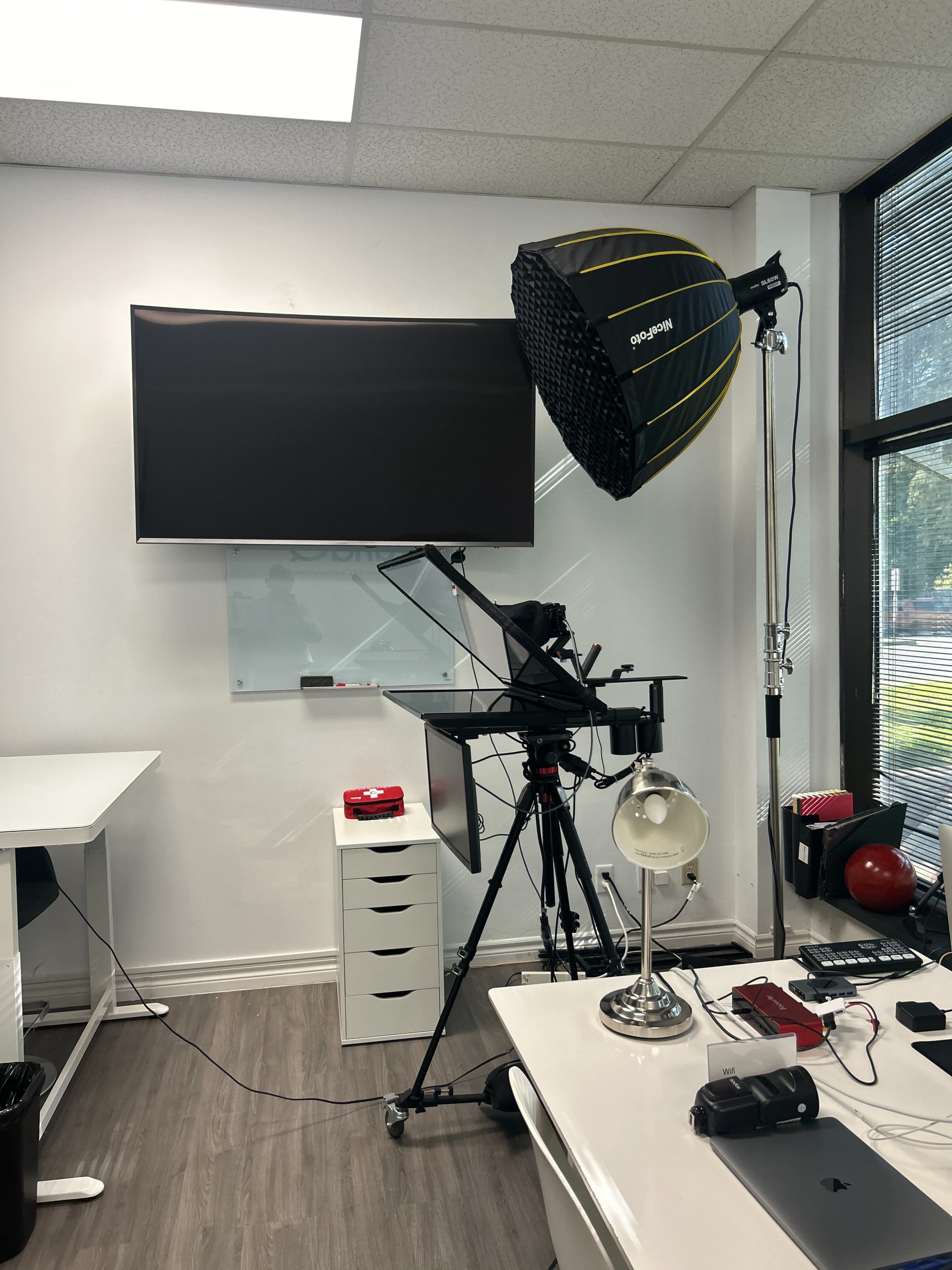 The image shows a bright office setup featuring a camera rig with lighting equipment, a wall-mounted TV, and a workspace with various electronic devices and accessories.