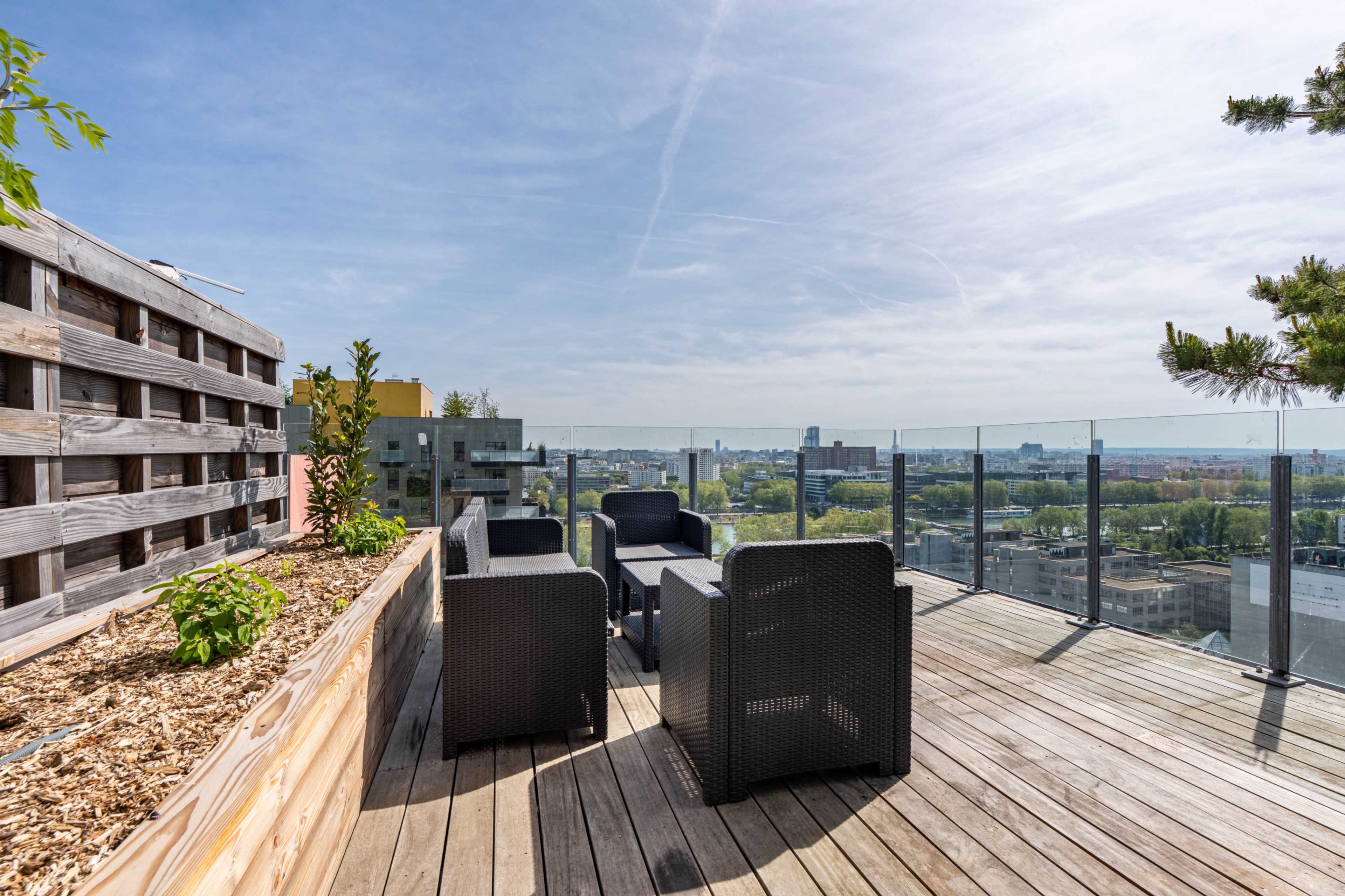 A spacious rooftop deck features black lounge chairs and a wooden planter, overlooking a cityscape under a clear sky.