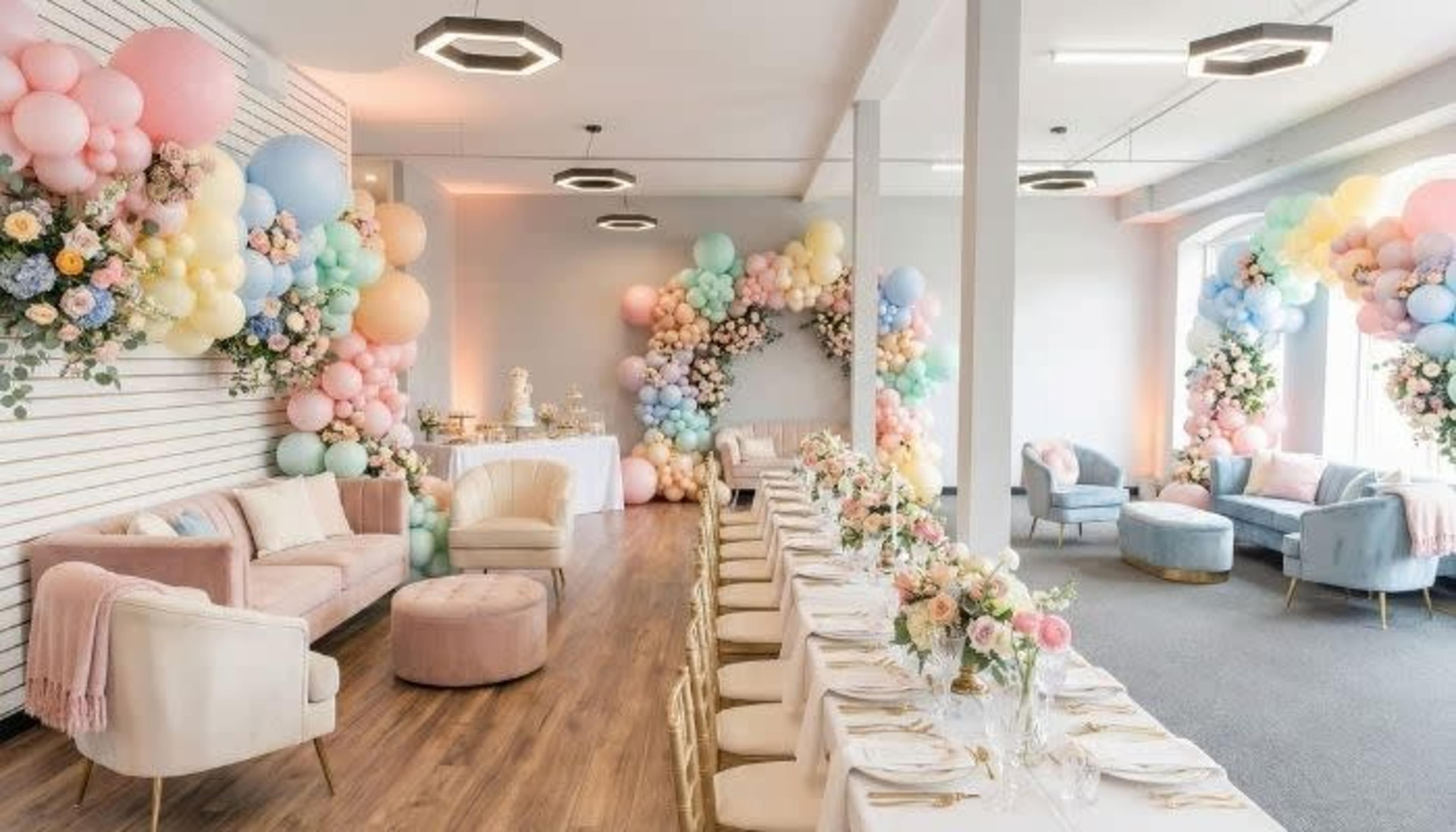 The image shows a decorated event space with elegant seating, a long dining table adorned with flowers, and colorful balloon arrangements creating a festive atmosphere.