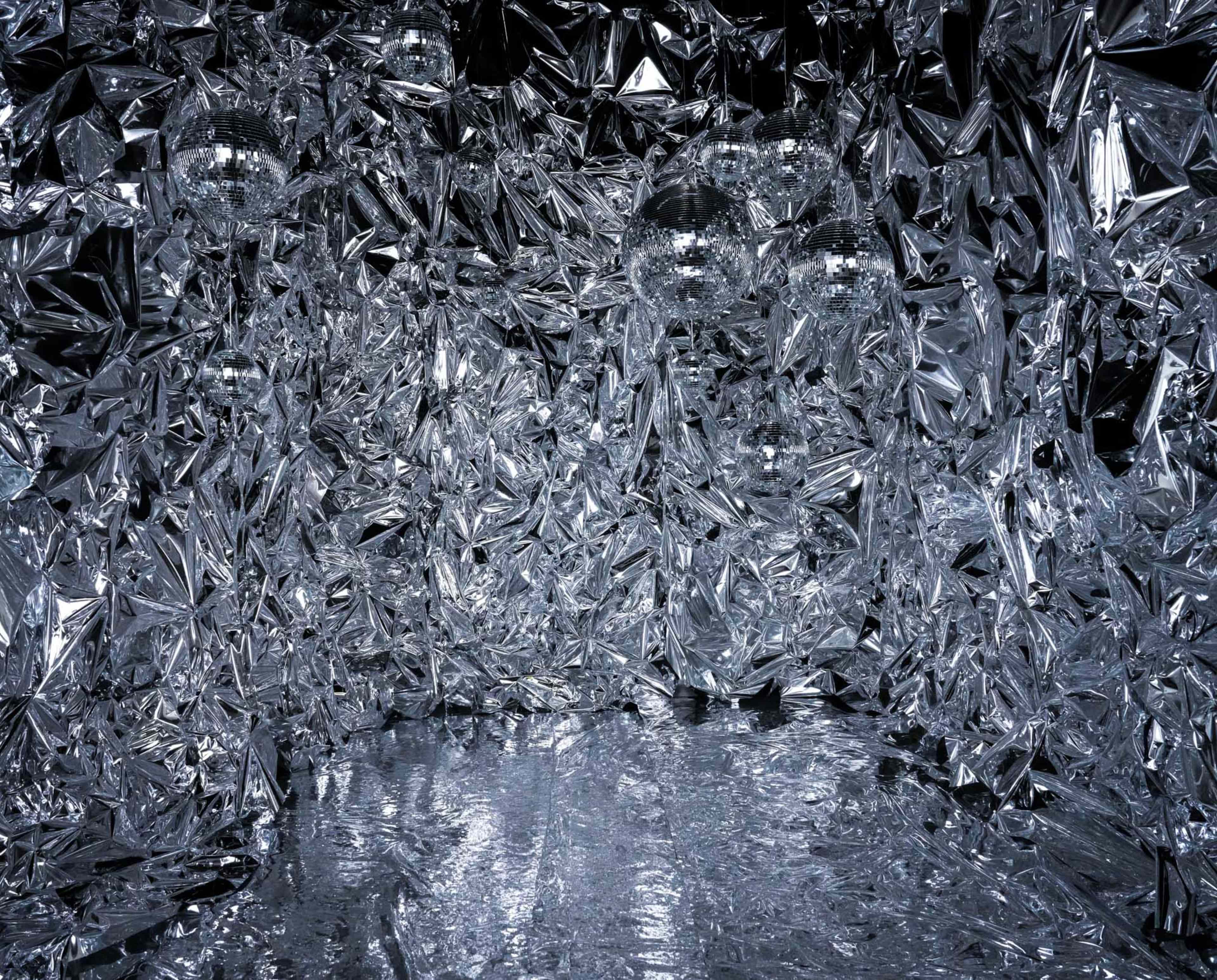 A room is covered with reflective, crumpled silver foil, creating an immersive, shiny environment with three disco balls hanging from the ceiling.