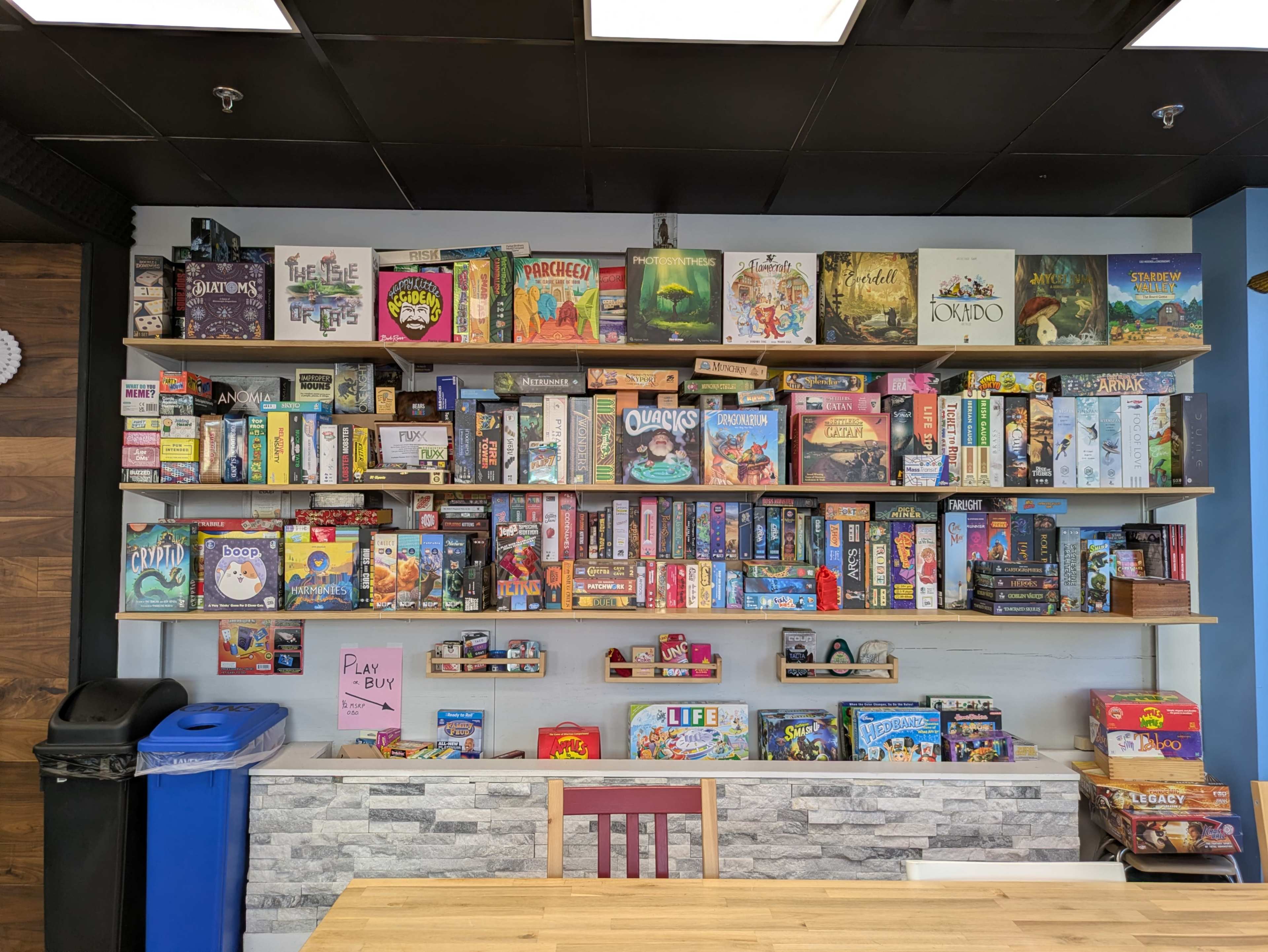 A wall-mounted shelf displays a variety of board games arranged by size and type, with a table and trash bin visible in the foreground.