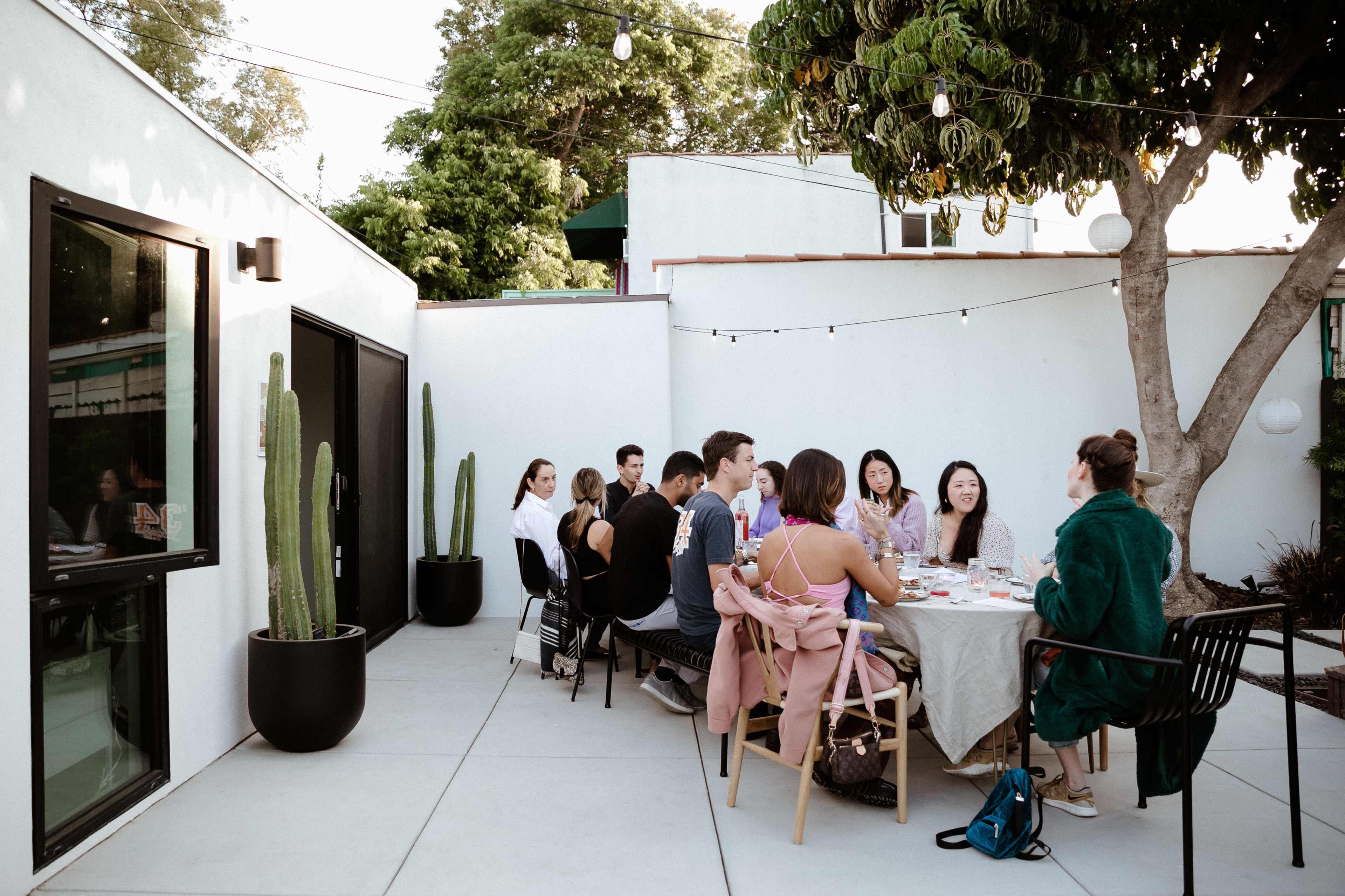 A group of people sits around a dining table in an outdoor setting with string lights and cacti in the background.