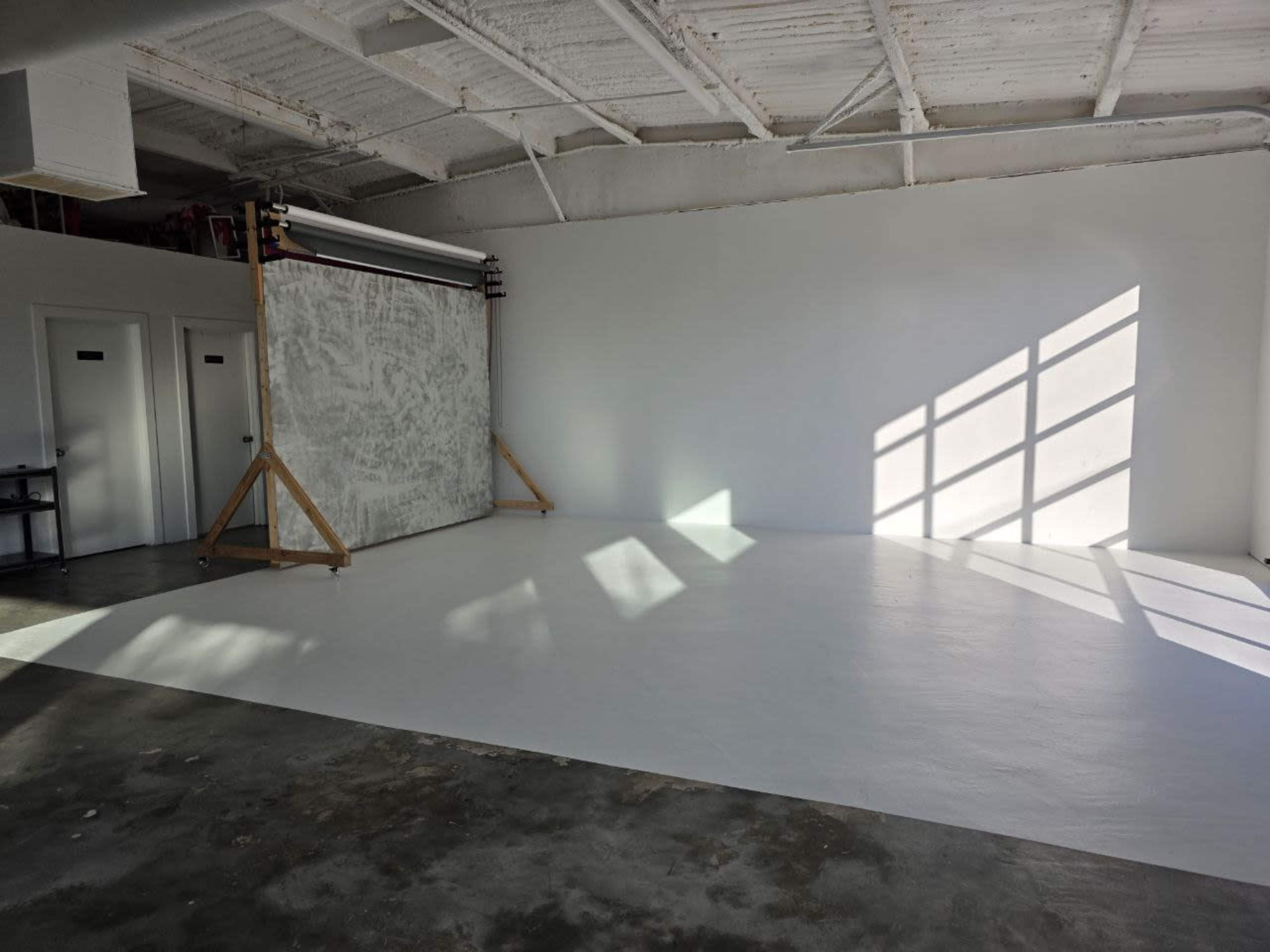 The image shows a spacious studio with a white floor, a large backdrop on a wooden frame, and sunlight casting shadows from a nearby window.