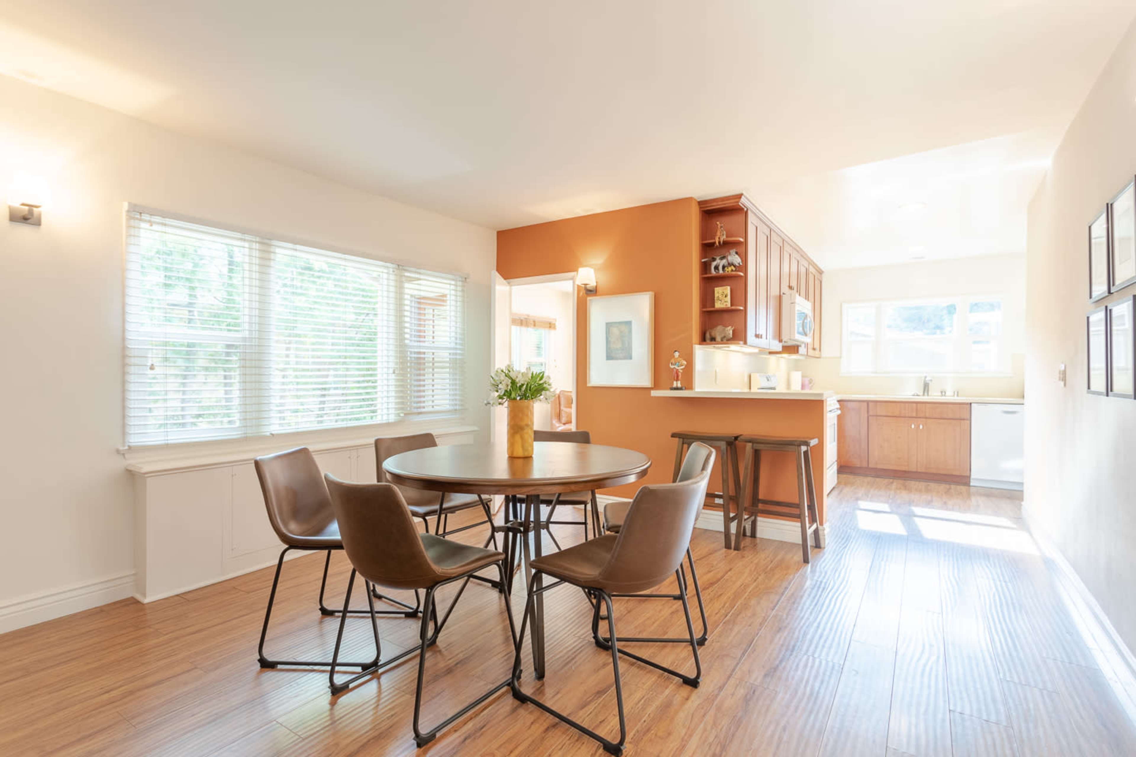 A dining area features a round table surrounded by four chairs, with a view of a bright kitchen and large windows.