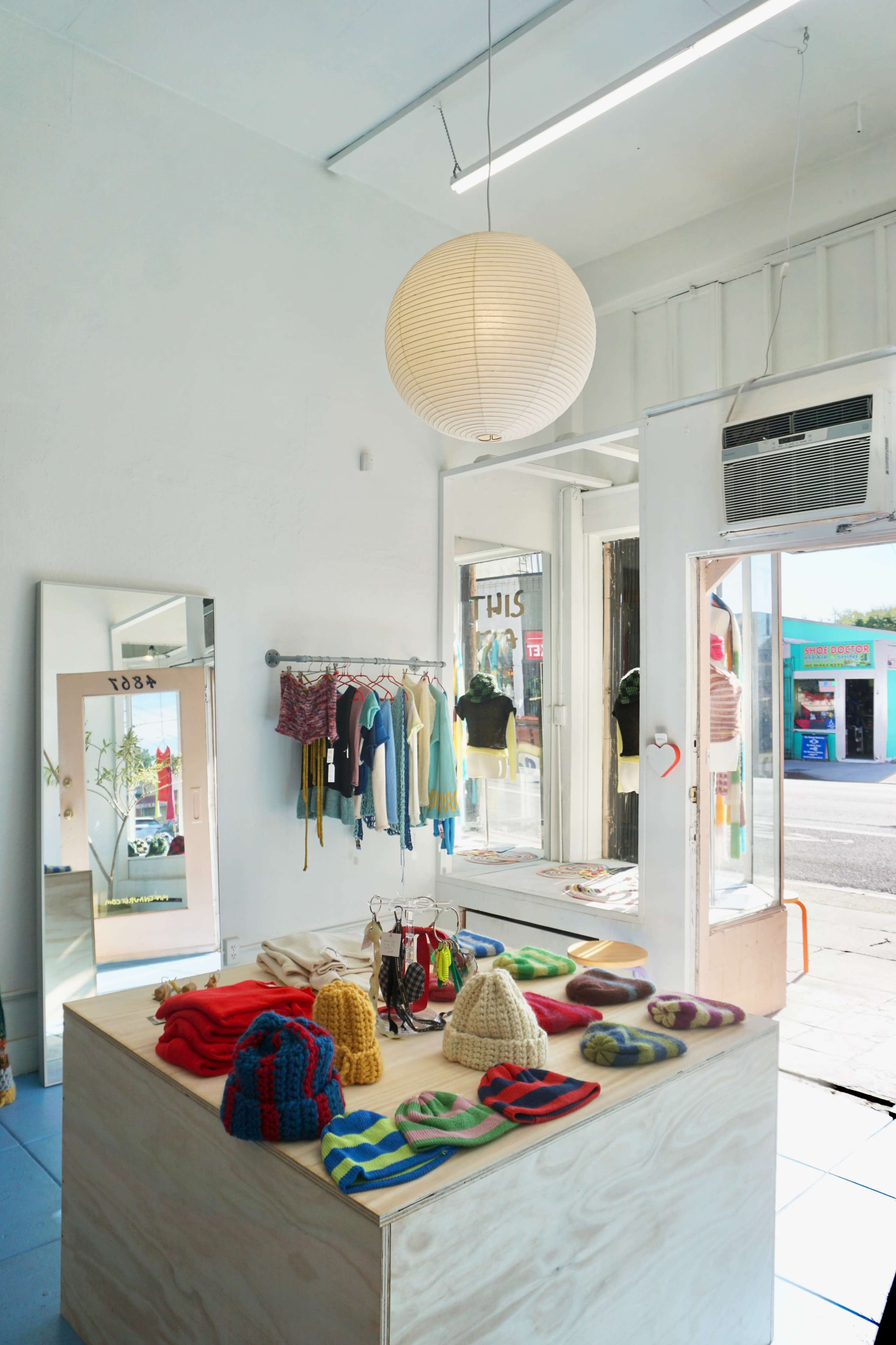 A brightly lit clothing store features a wooden table displaying colorful knitted hats and accessories, with clothing racks and a large mirror in the background.
