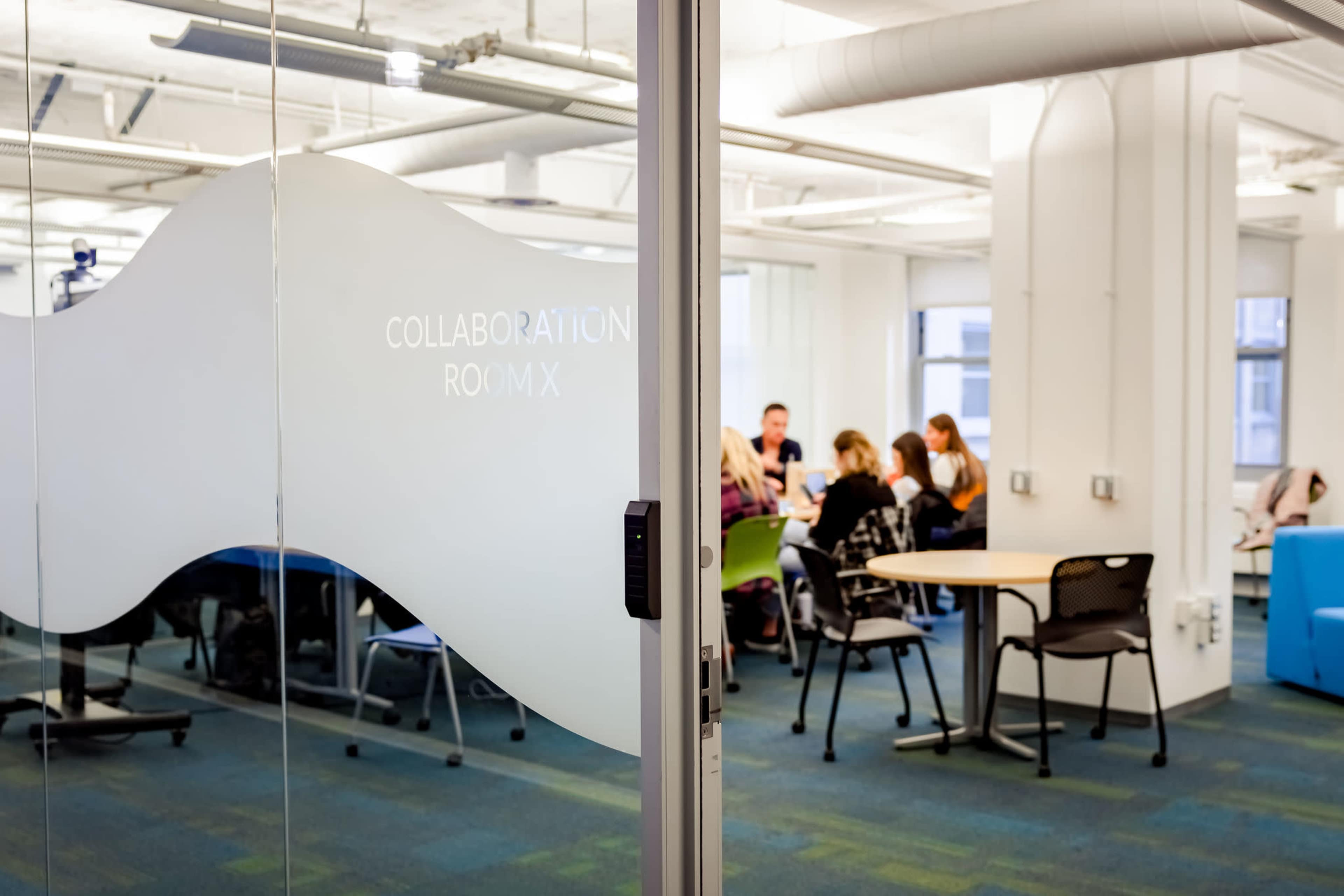 A glass door leading to a collaboration room where a group of people is seated around a table.