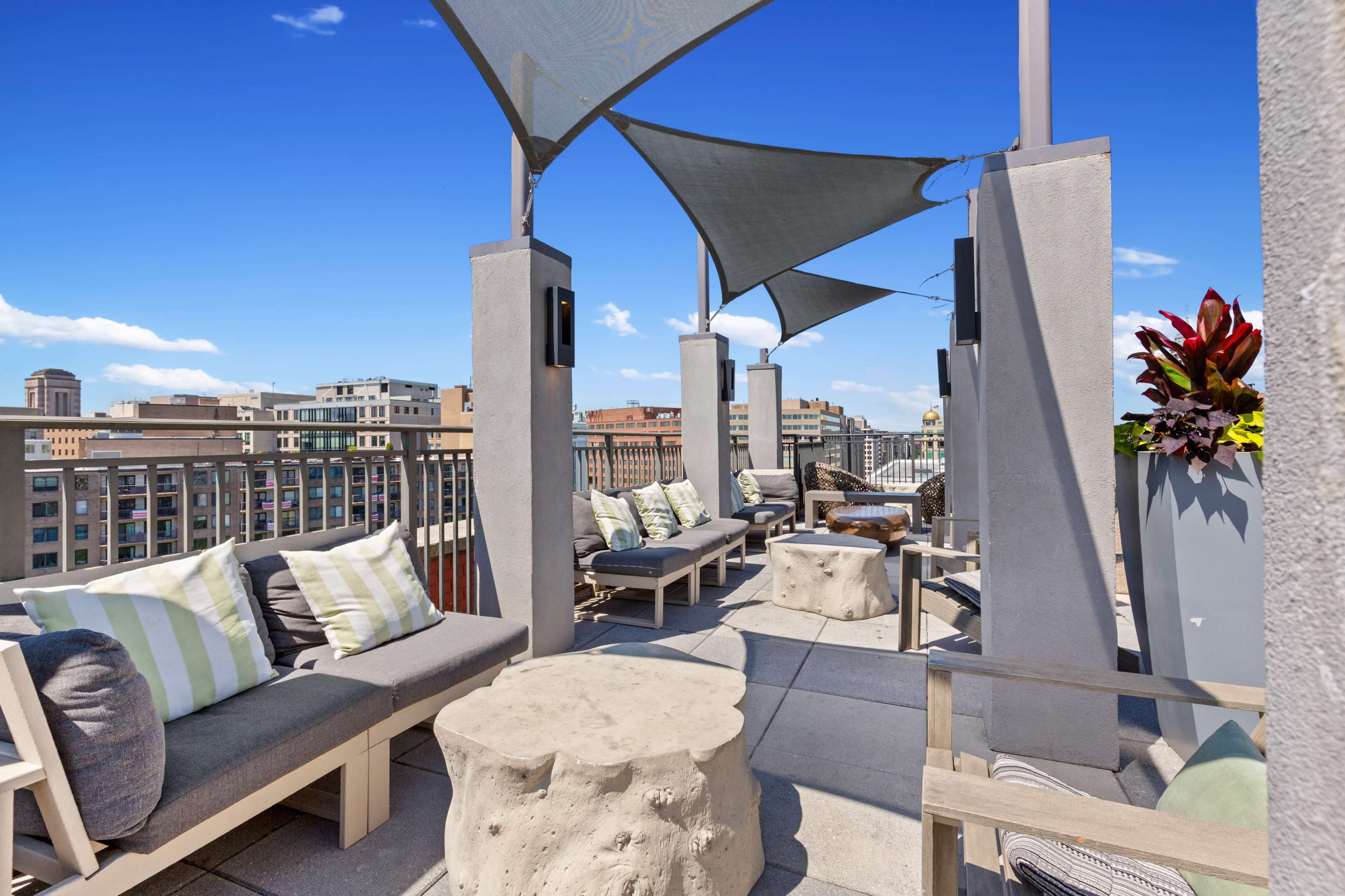 The image shows a rooftop terrace featuring sofas with cushions, stone tables, and shade sails, overlooking a city skyline.