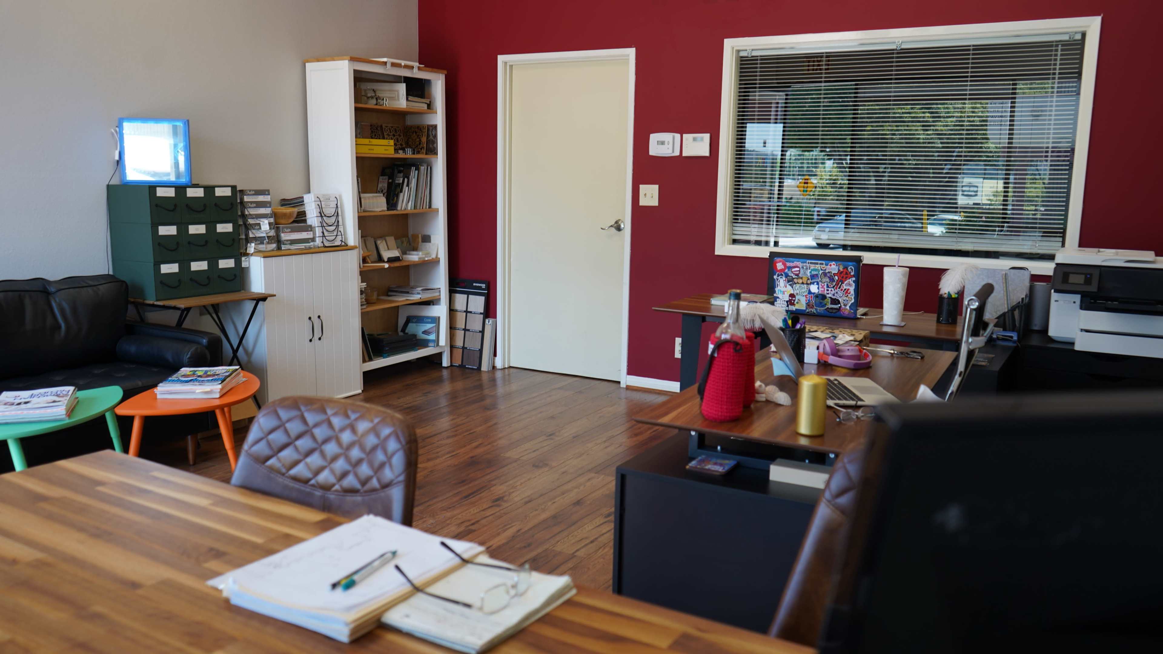 The image depicts a modern office space with wooden flooring, a red accent wall, and furniture including a desk, chairs, shelves, and a printer.