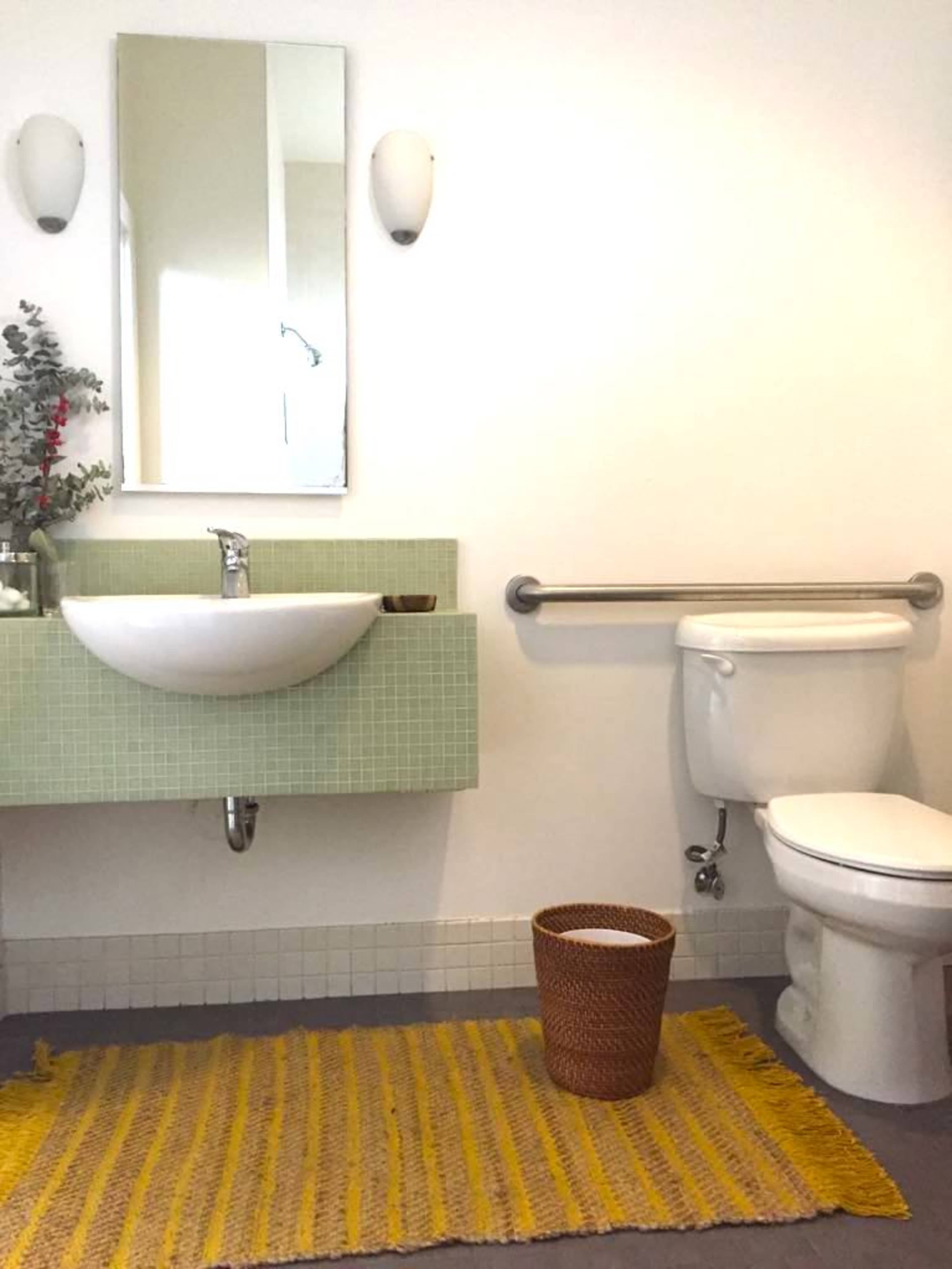 A modern bathroom features a wall-mounted sink, a white toilet, and a woven basket on a yellow rug.